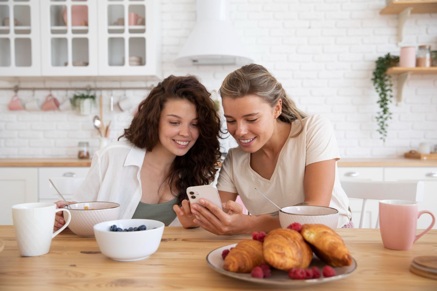 Two women smiling at a phone in a light kitchen; breakfast food and cups on the table.