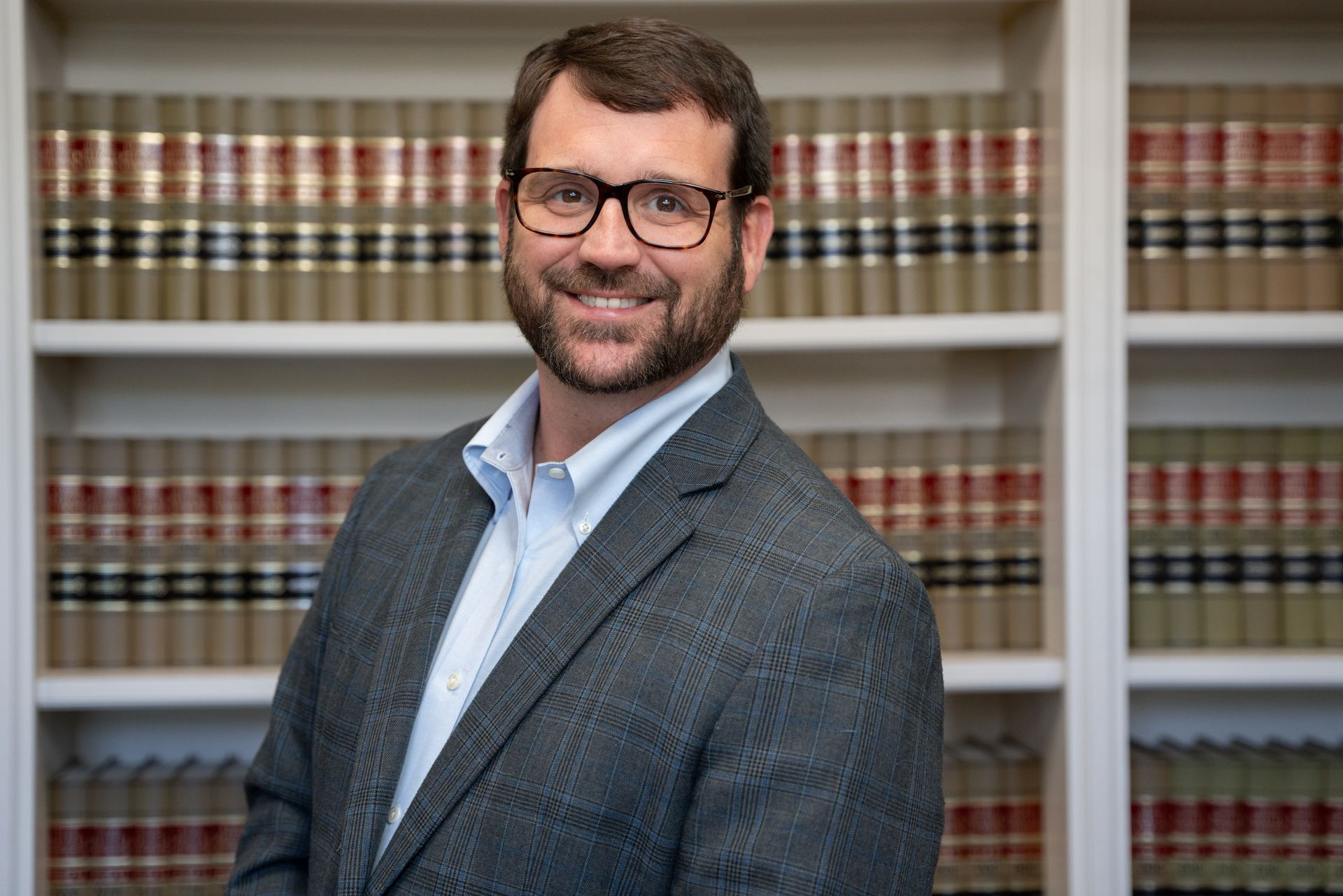 Man in glasses and suit smiles in front of a bookshelf.