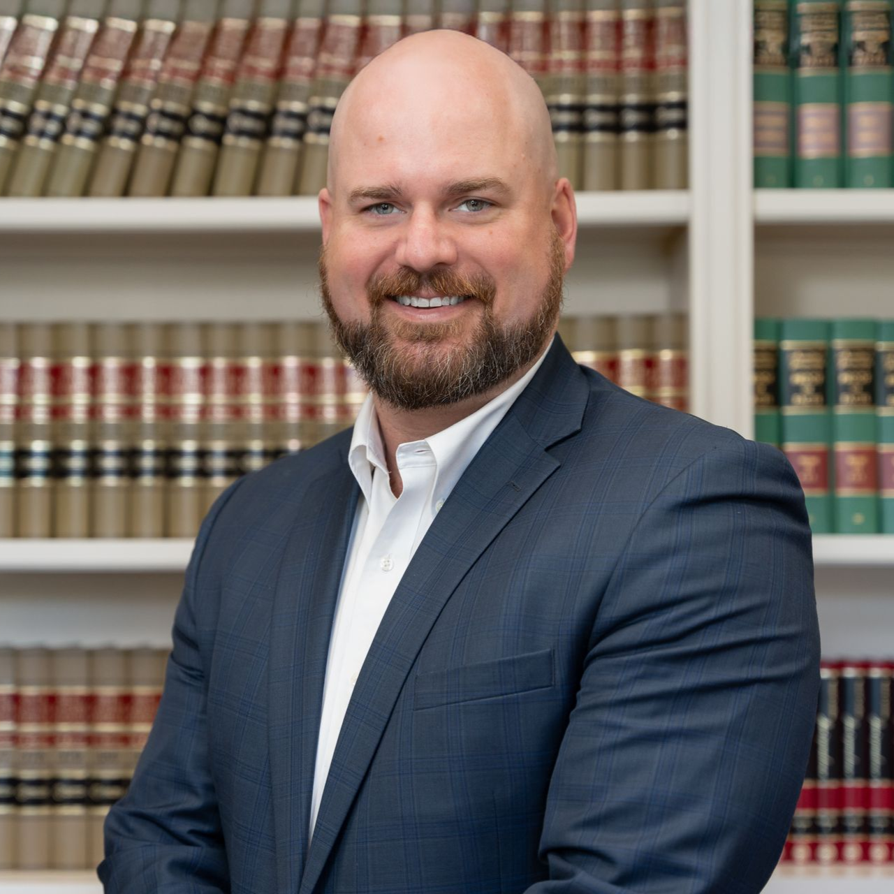 Bald man with a beard in a suit, smiling in front of a bookshelf.