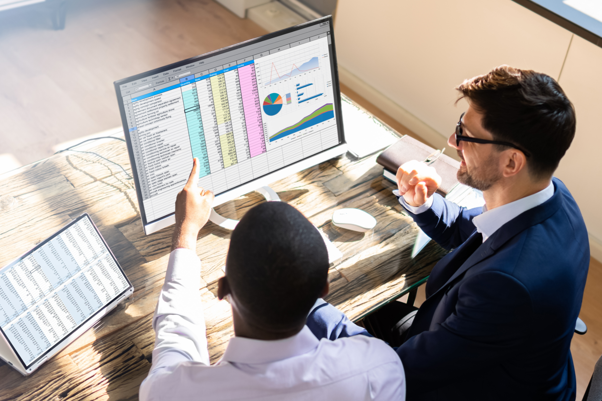 Two men in a bright office, examining financial data on a computer screen. One points, the other holds a pen.