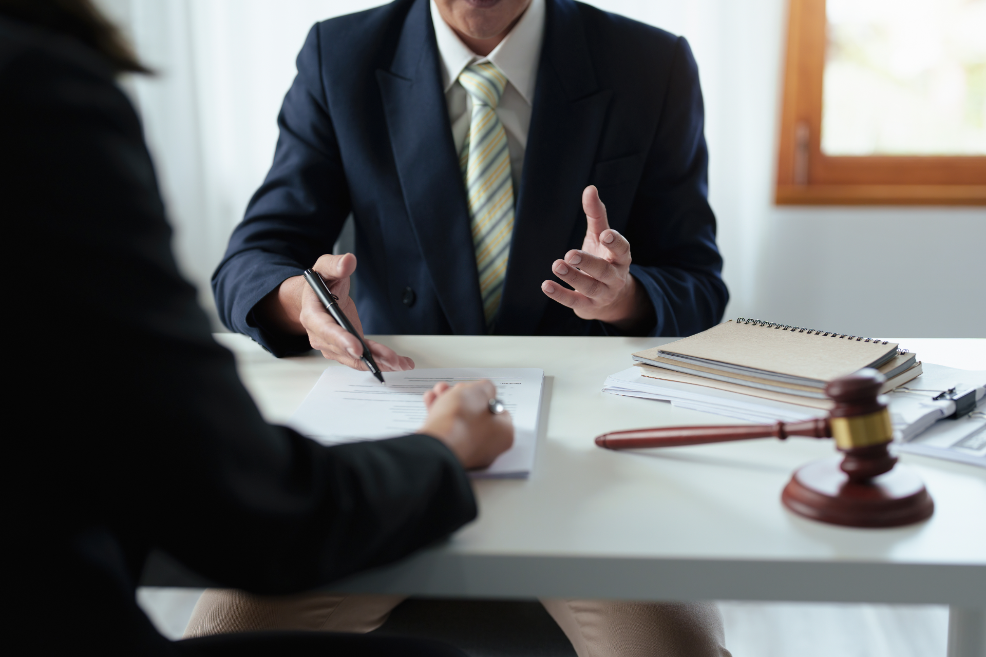 Lawyer in suit gestures, discussing documents with a client at a table, gavel present.