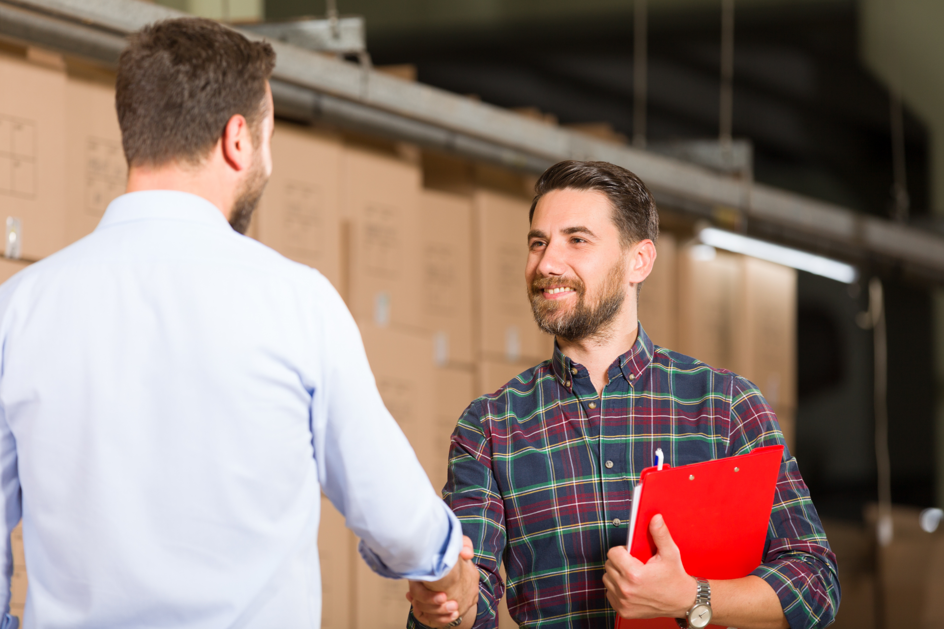 Two men shaking hands in a warehouse, one holding a red clipboard, boxes in background.