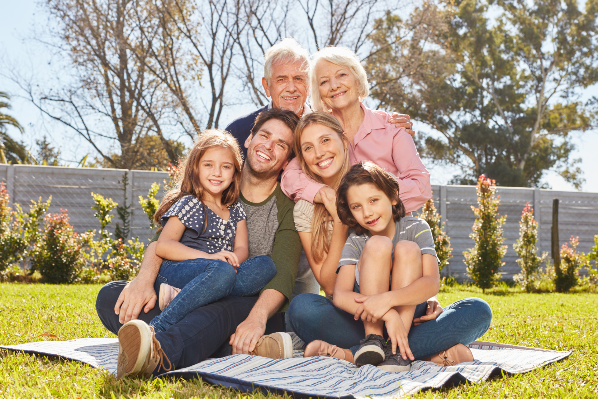 Family of seven smiling together on a picnic blanket in a sunny backyard.