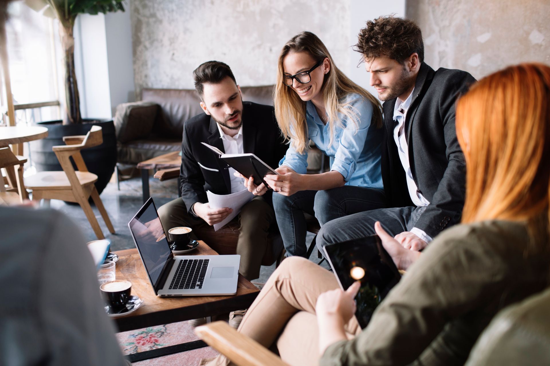 Group of people collaborating in a cafe; reviewing notes and using laptops and tablets.