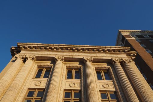 Beige building facade with columns, beneath a clear blue sky, adjacent to a brown brick building.