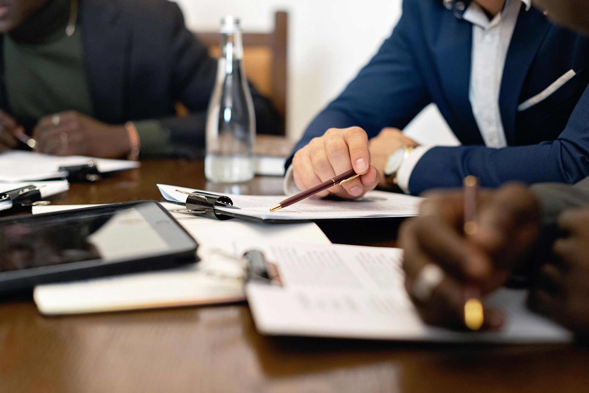 People in business suits review documents at a table, with pens and a tablet visible.