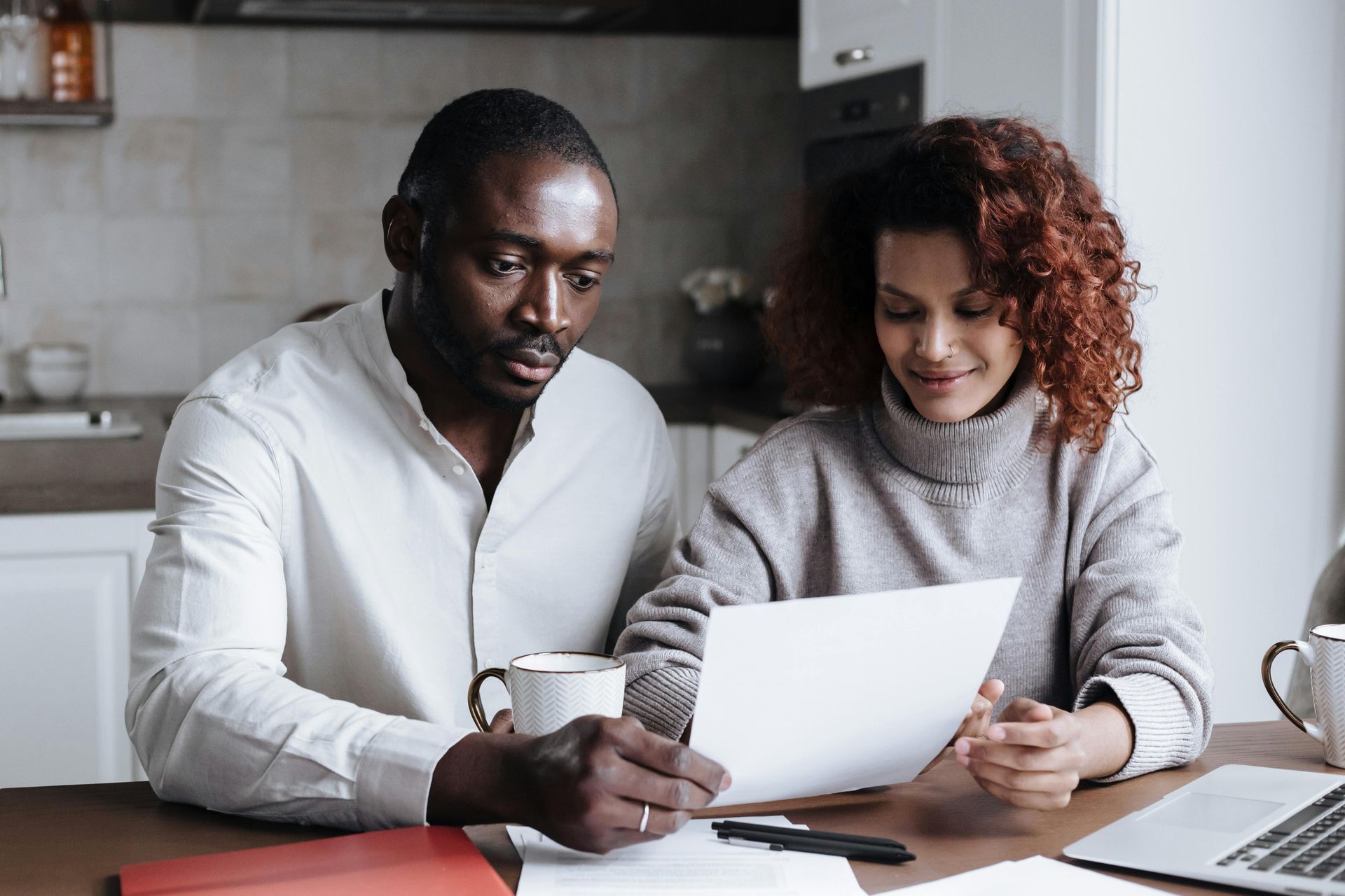 Couple reviewing documents at a table. Man in white shirt, woman in sweater, coffee mugs, laptop.