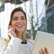 A woman is smiling while holding a tablet and talking on a cell phone