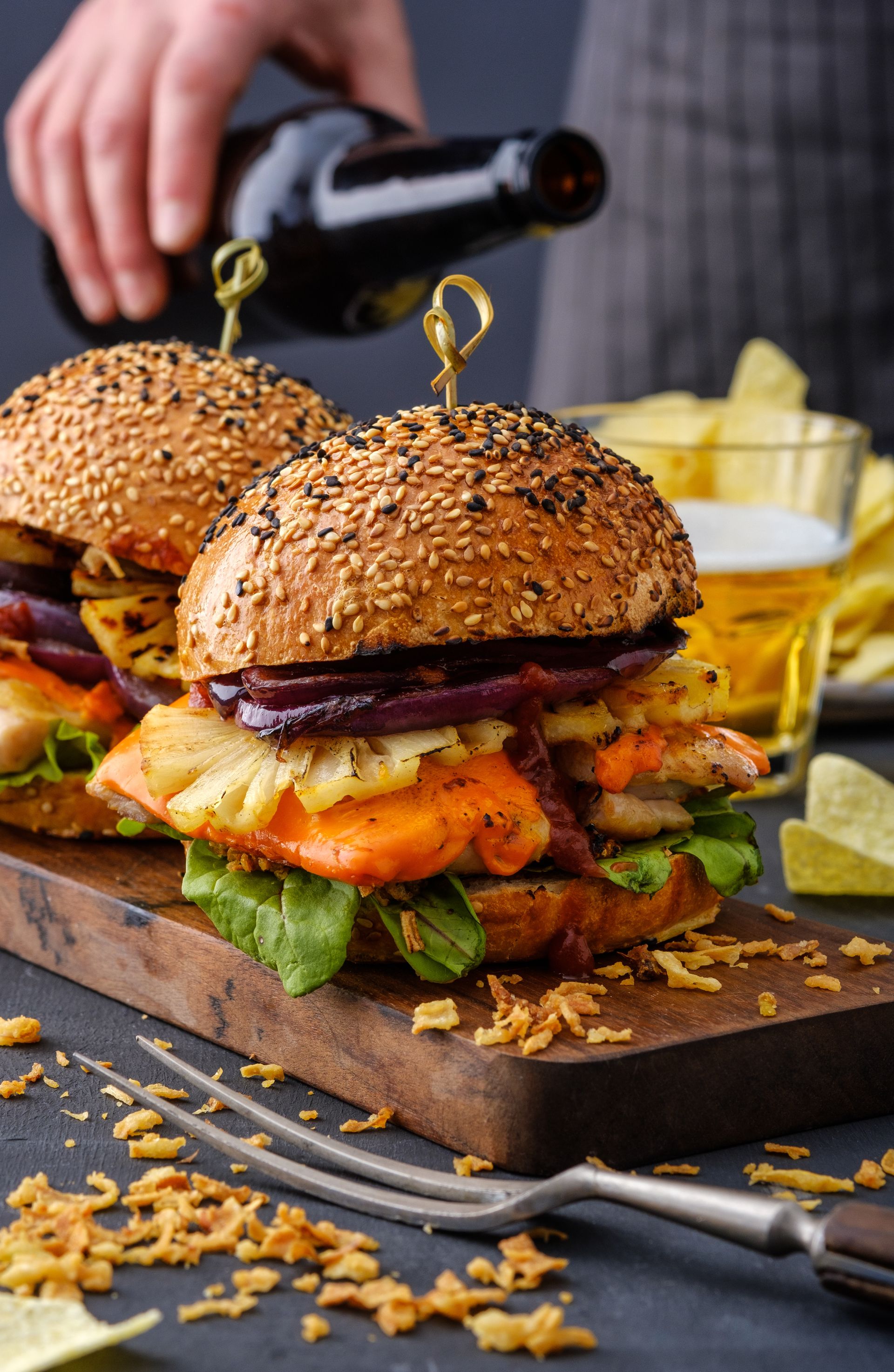 A close-up shot of two gourmet burgers served on a wooden board, featuring sesame seed buns, melted orange cheese, grilled pineapple rings, and red onions. In the background, a person pours beer from a dark glass bottle, alongside a side of potato chips and scattered crispy fried onions on a dark tabletop.