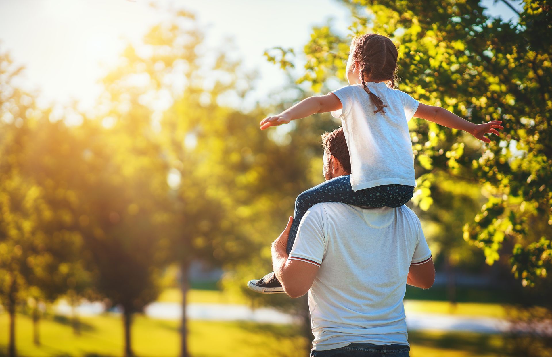 A view from behind of a father carrying his young daughter on his shoulders while walking through a park at sunset. The little girl has her arms spread wide out to the sides, facing toward the warm, bright sunlight filtering through the green trees.