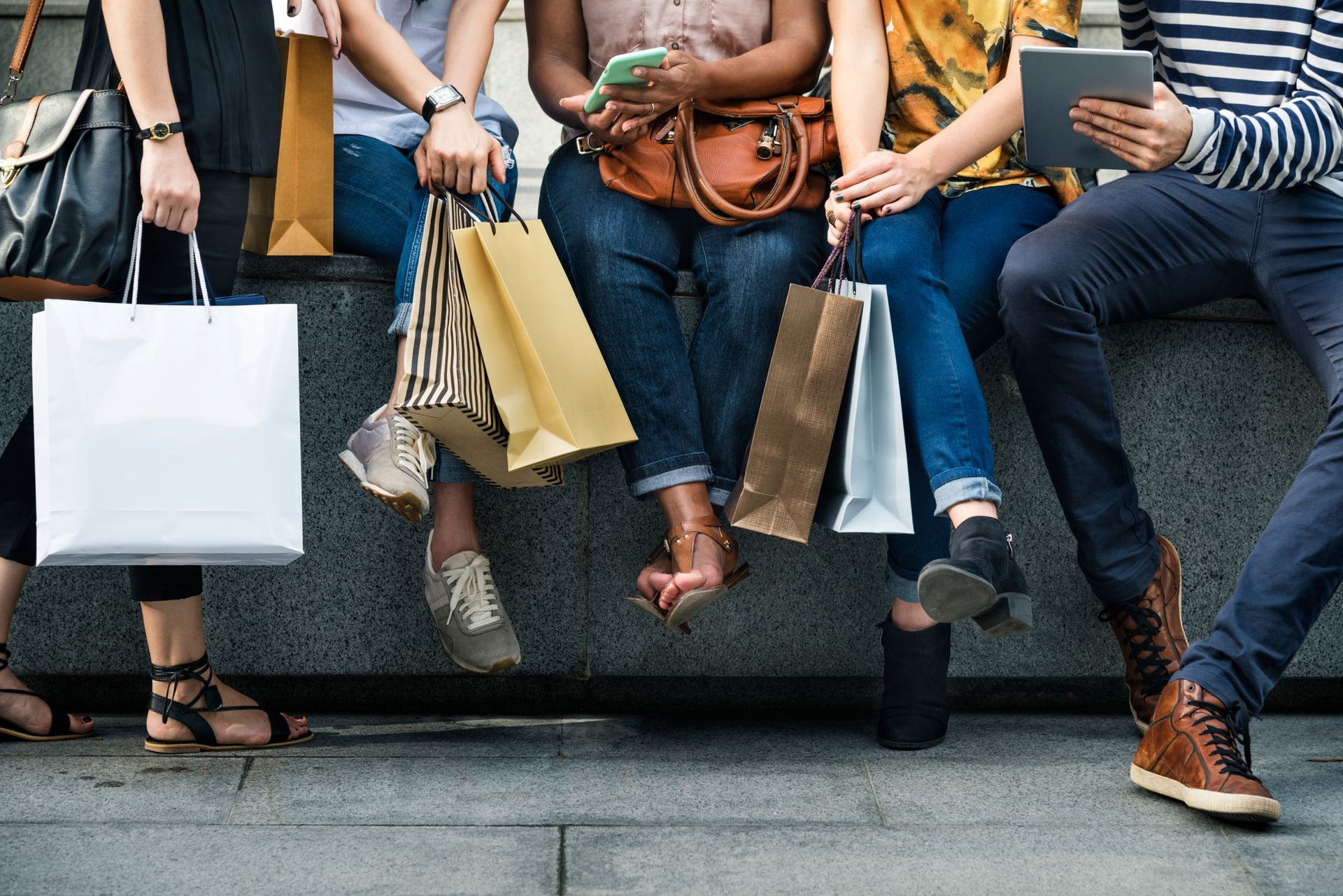 A cropped, low-angle shot of a group of people sitting side-by-side on an outdoor ledge after a shopping trip. The focus is on their legs and hands as they hold various white, brown, and gold paper shopping bags, while some look at a smartphone and a digital tablet.