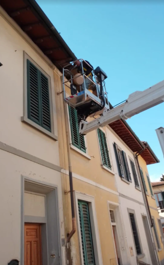 Una piattaforma elevatrice tiene un operaio vicino al tetto di un edificio per le riparazioni; persiane e cielo blu, porta color pesca.