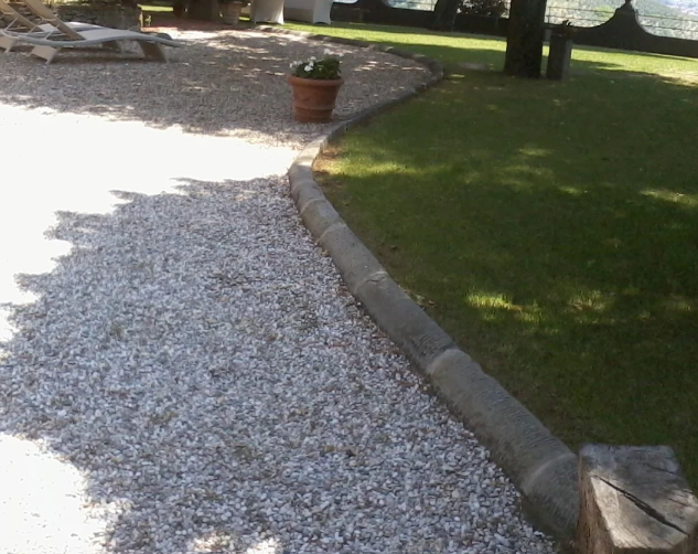 Sentiero di ghiaia che costeggia il prato verde, separato da un cordolo di cemento. Nelle vicinanze c'è una pianta in vaso.