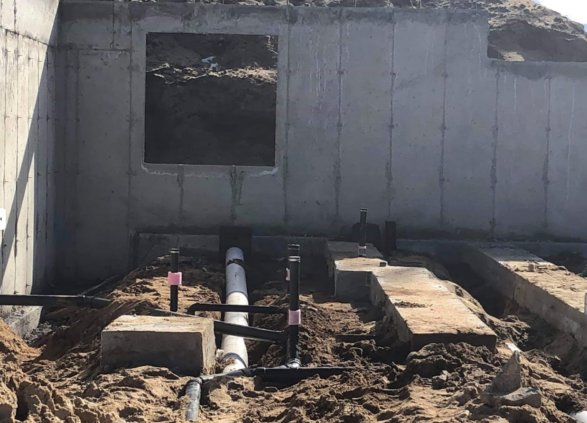 A concrete wall with a window and pipes in the dirt.