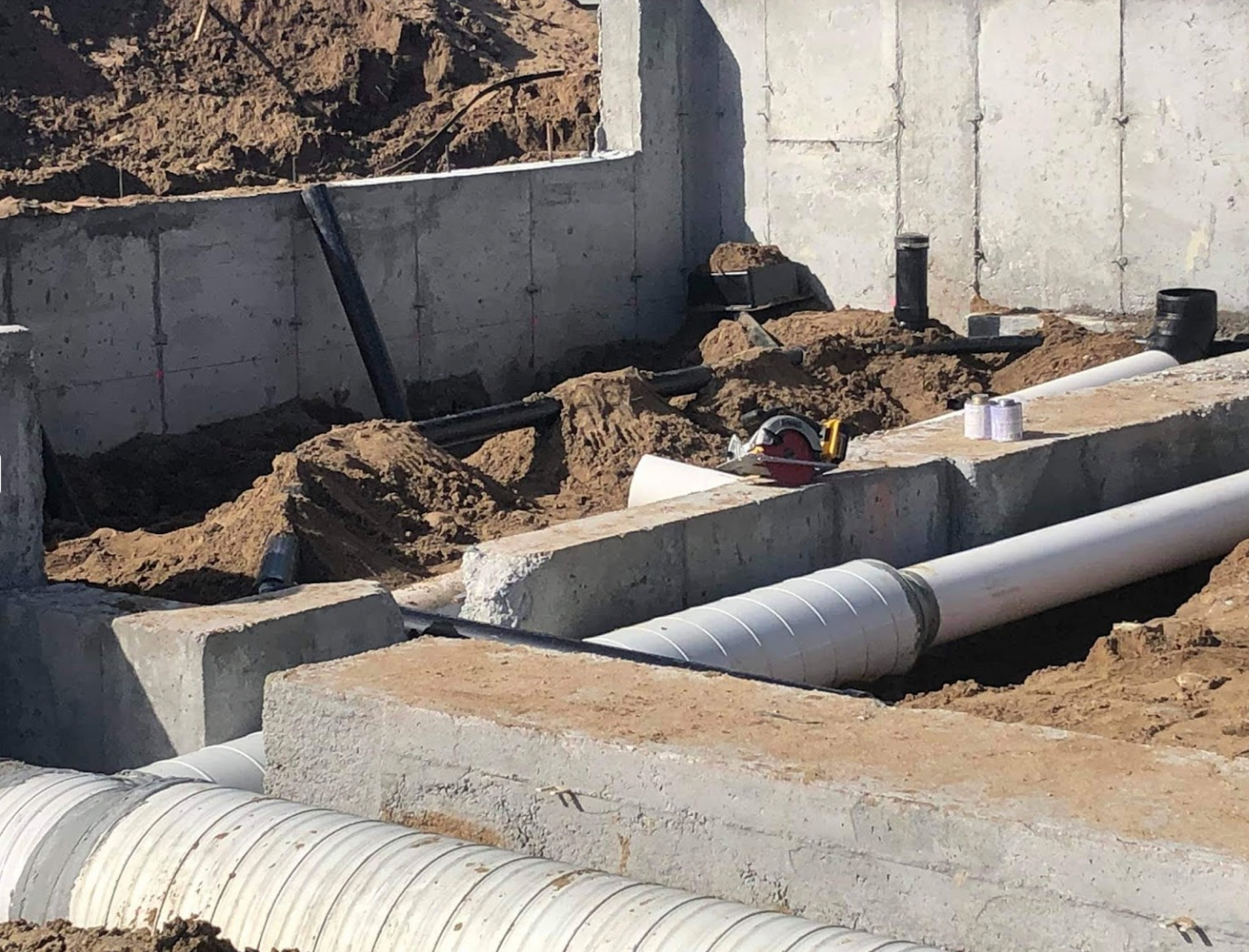 A bunch of pipes are laying in the dirt near a concrete wall