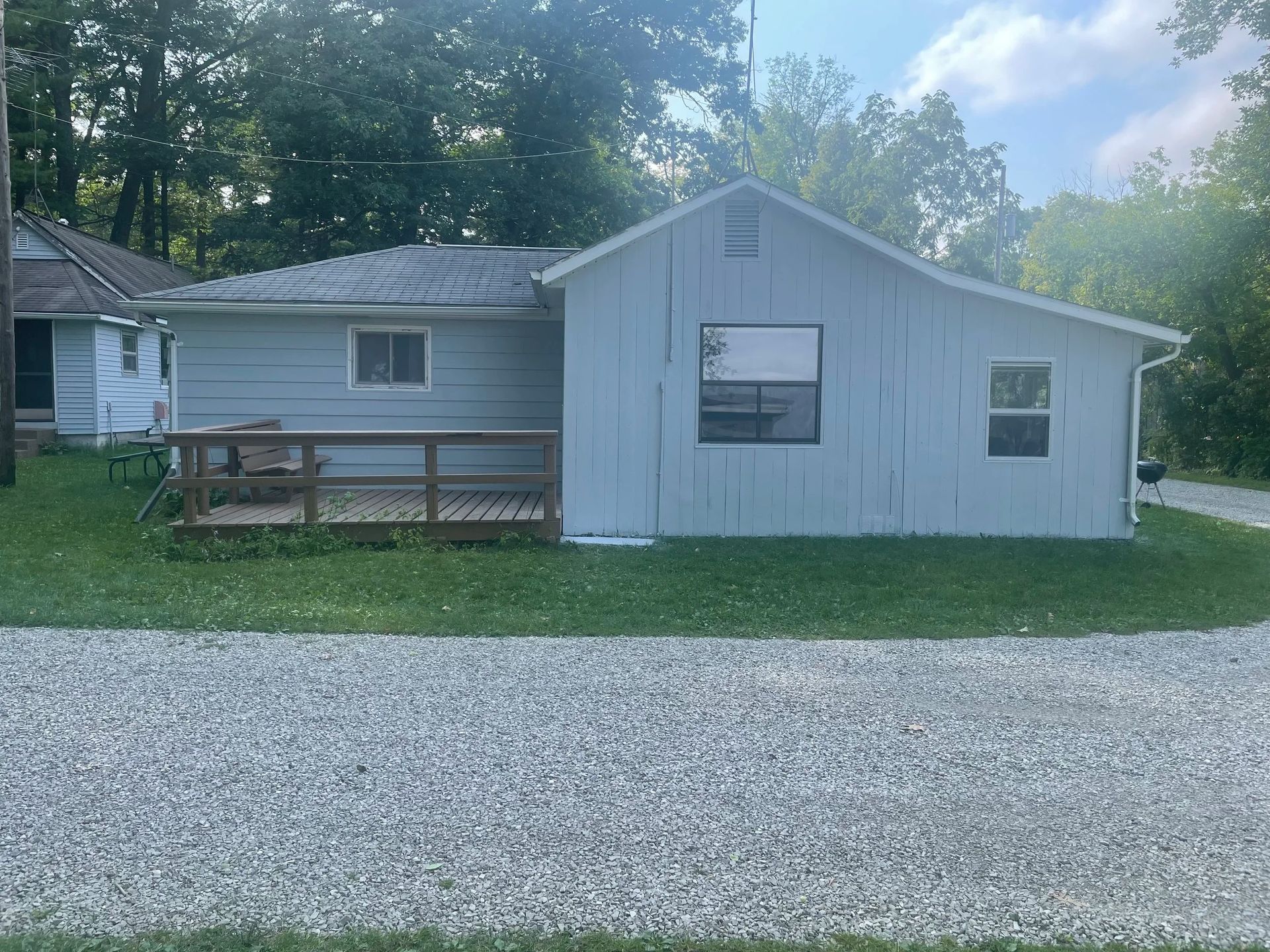 Small, light-blue cottage with windows and a gray patio in front of trees.