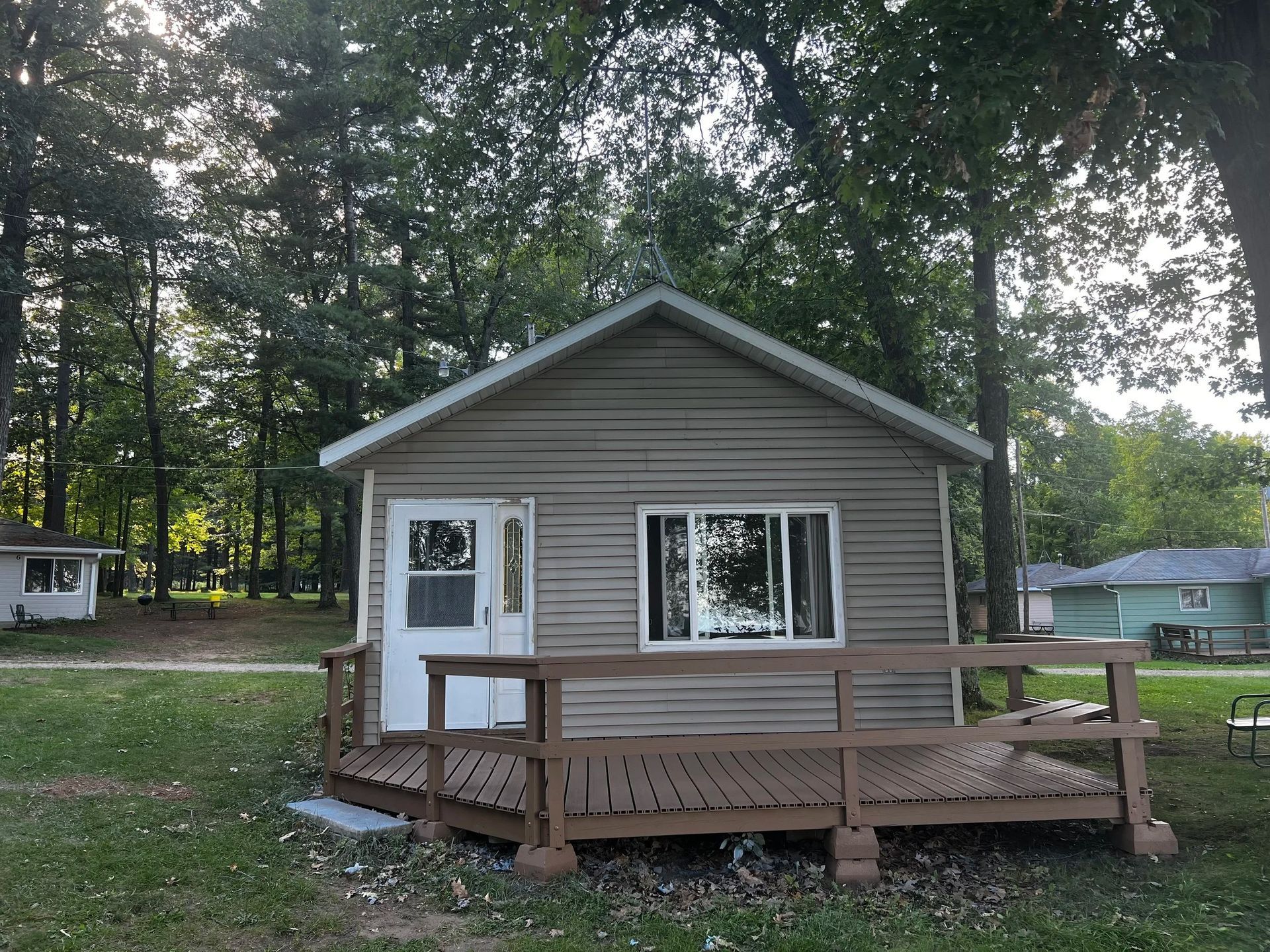 Small, weathered cabin with a brown deck. Trees surround the building, set in a grassy yard.