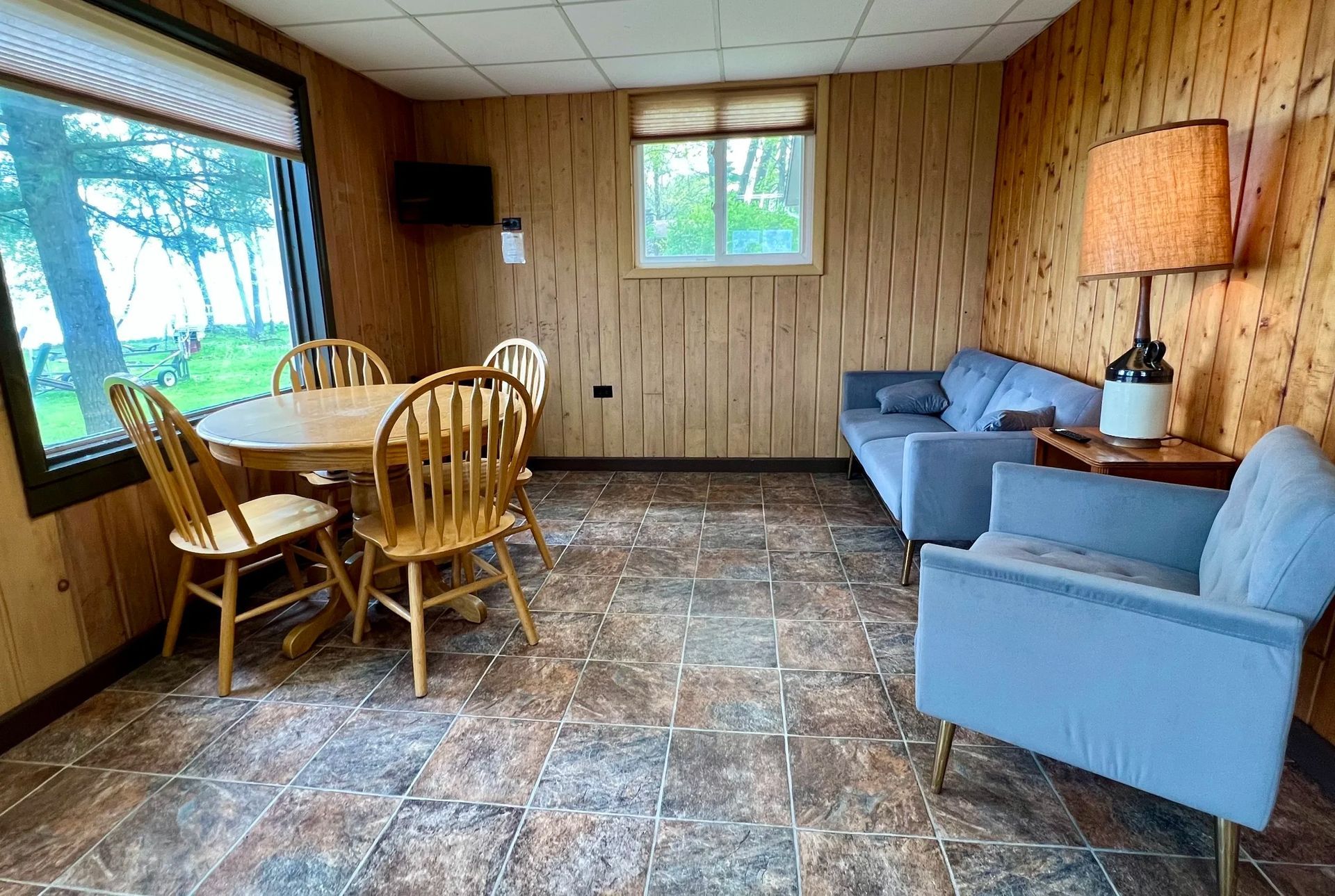 Small, light-blue cottage with windows and a gray patio in front of trees.