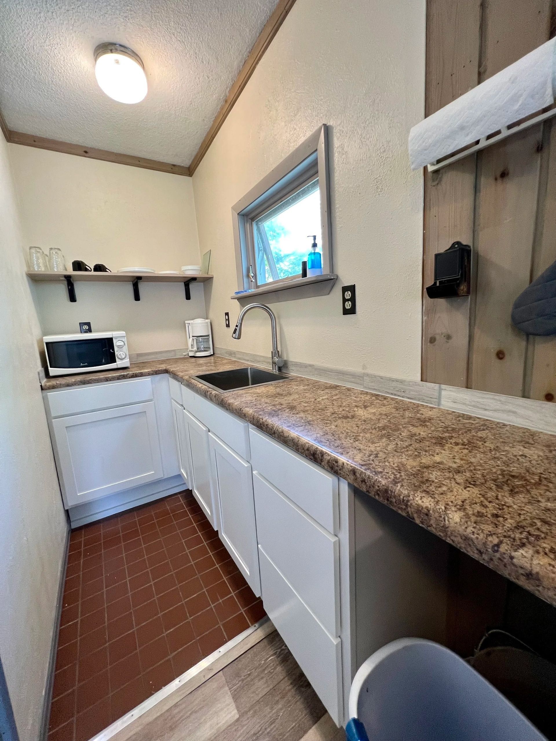 Small kitchenette with white cabinets, brown countertop, sink, microwave, window.