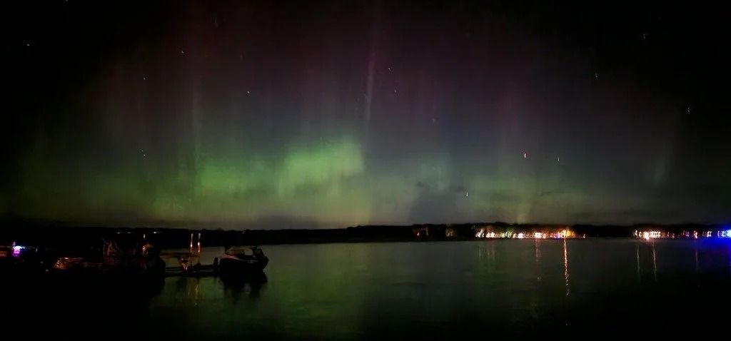 Green and purple aurora borealis over a dark lake with reflections of lights.