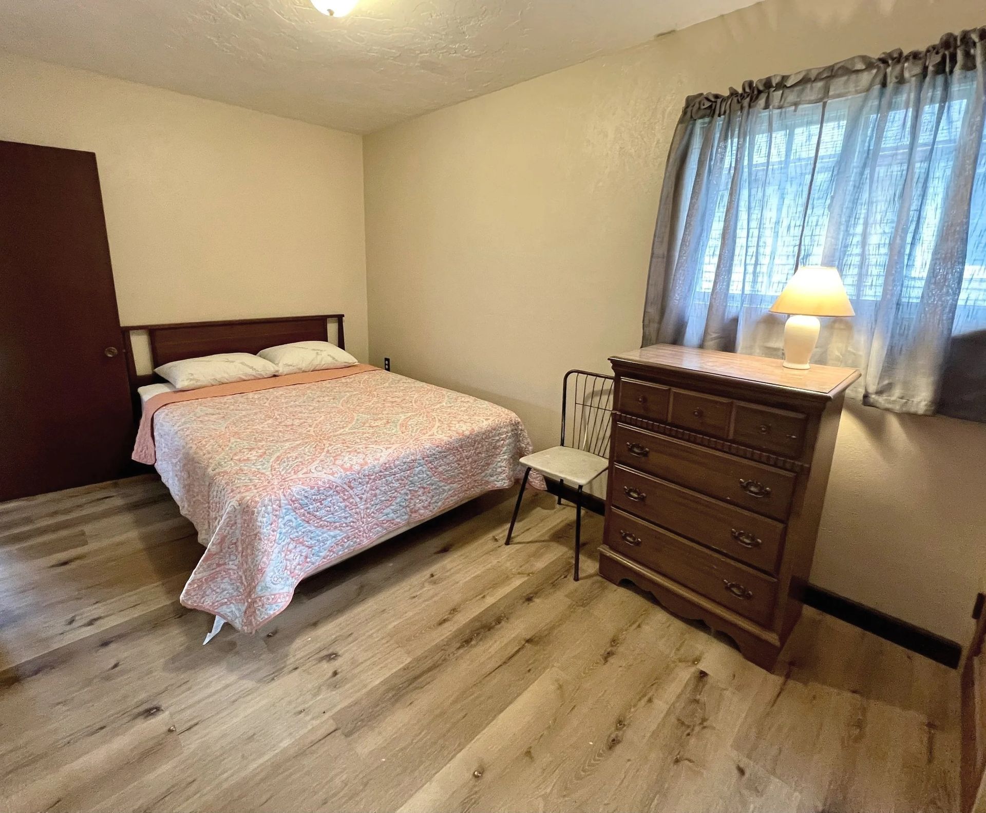 Bedroom with bed, dresser, window with curtains, and wood flooring.