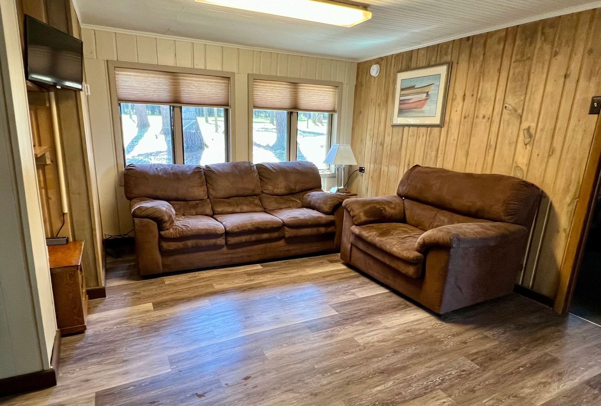 Living room with brown sofa and armchair, two windows with shades, wooden paneling, and a small TV.