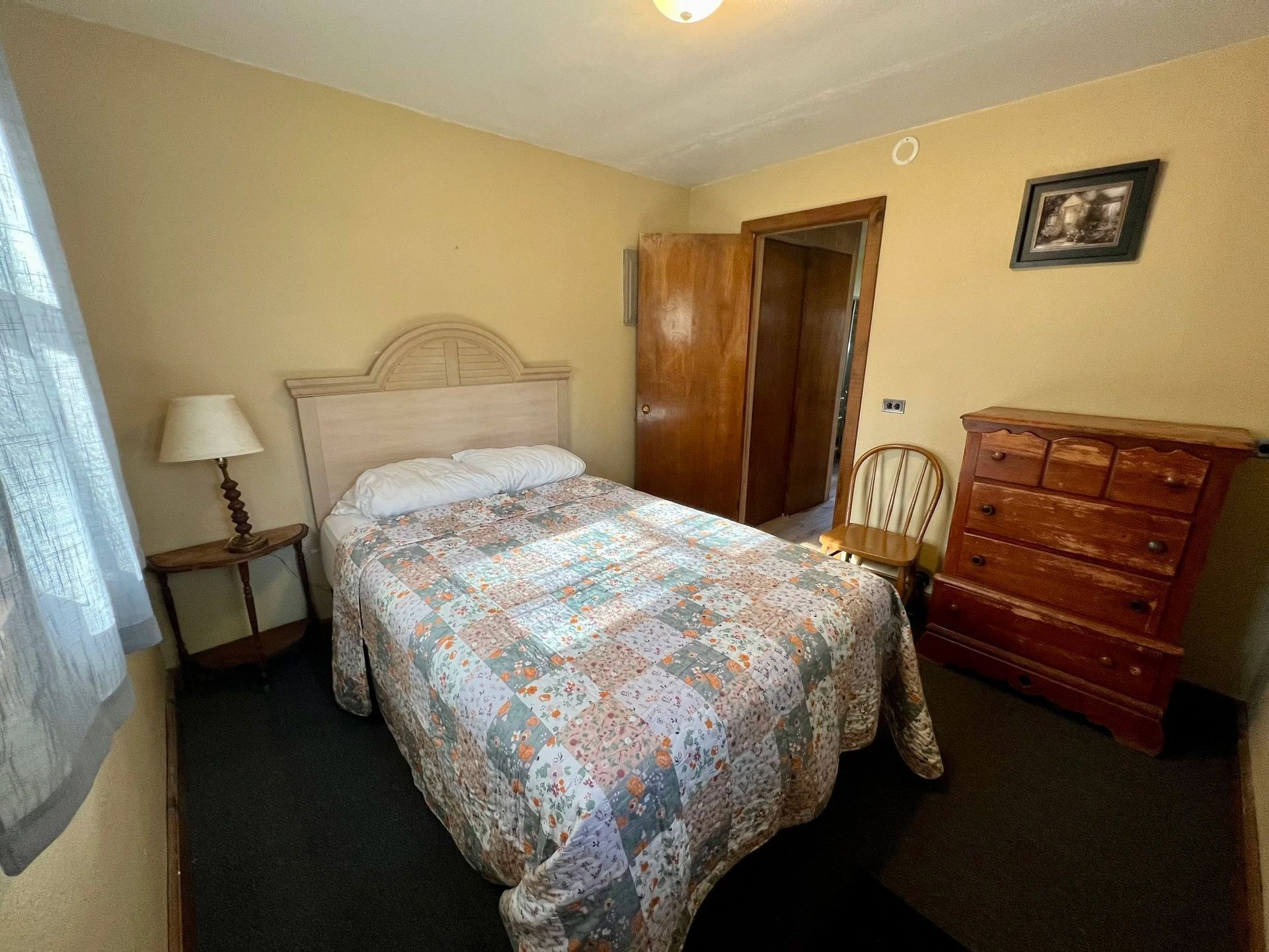 Bedroom with bed, nightstand, dresser, and open doorway. Yellow walls, floral quilt, dark carpet.