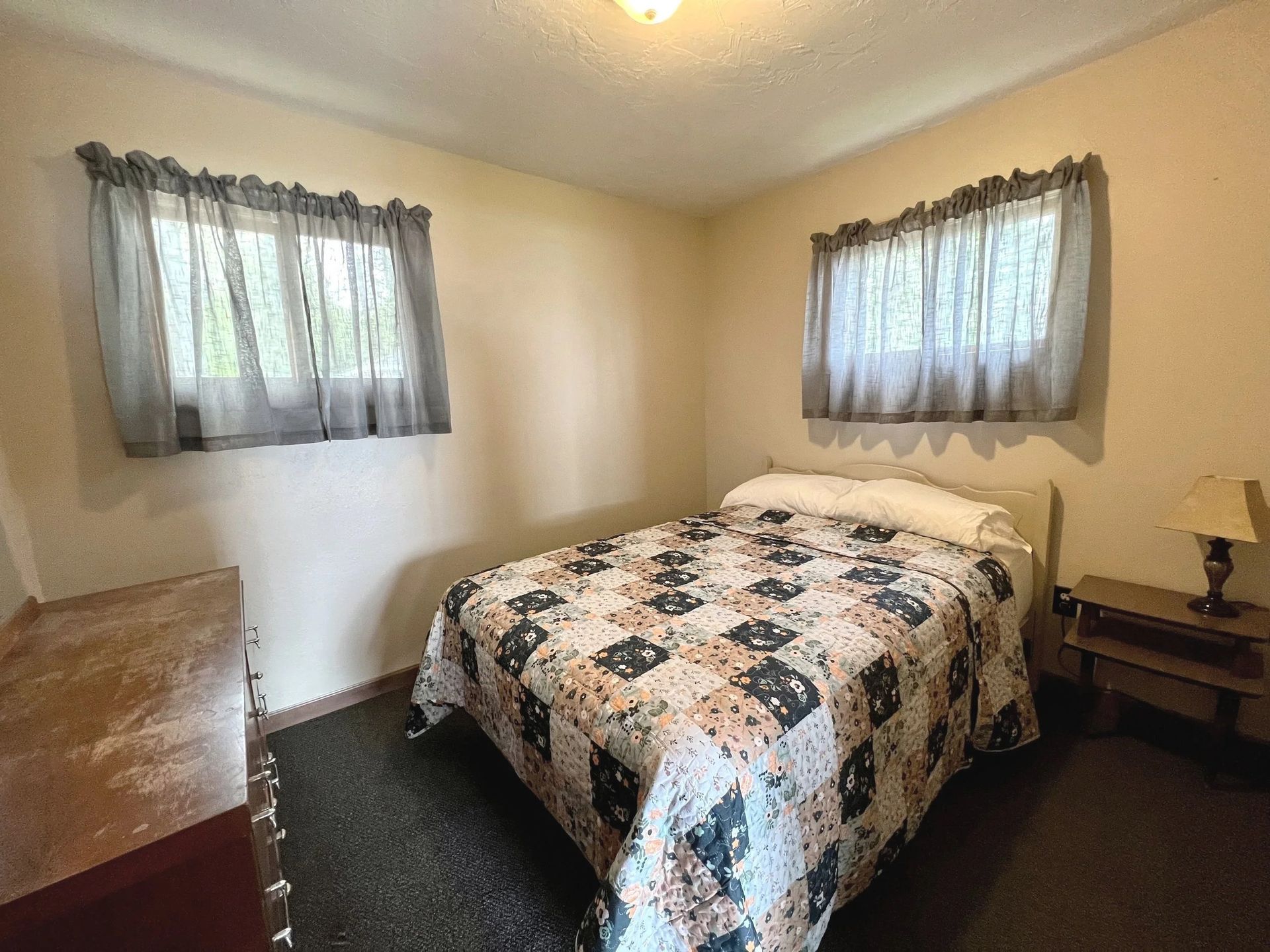 Bedroom with a bed, dresser, nightstand, two windows with gray curtains, and carpet.