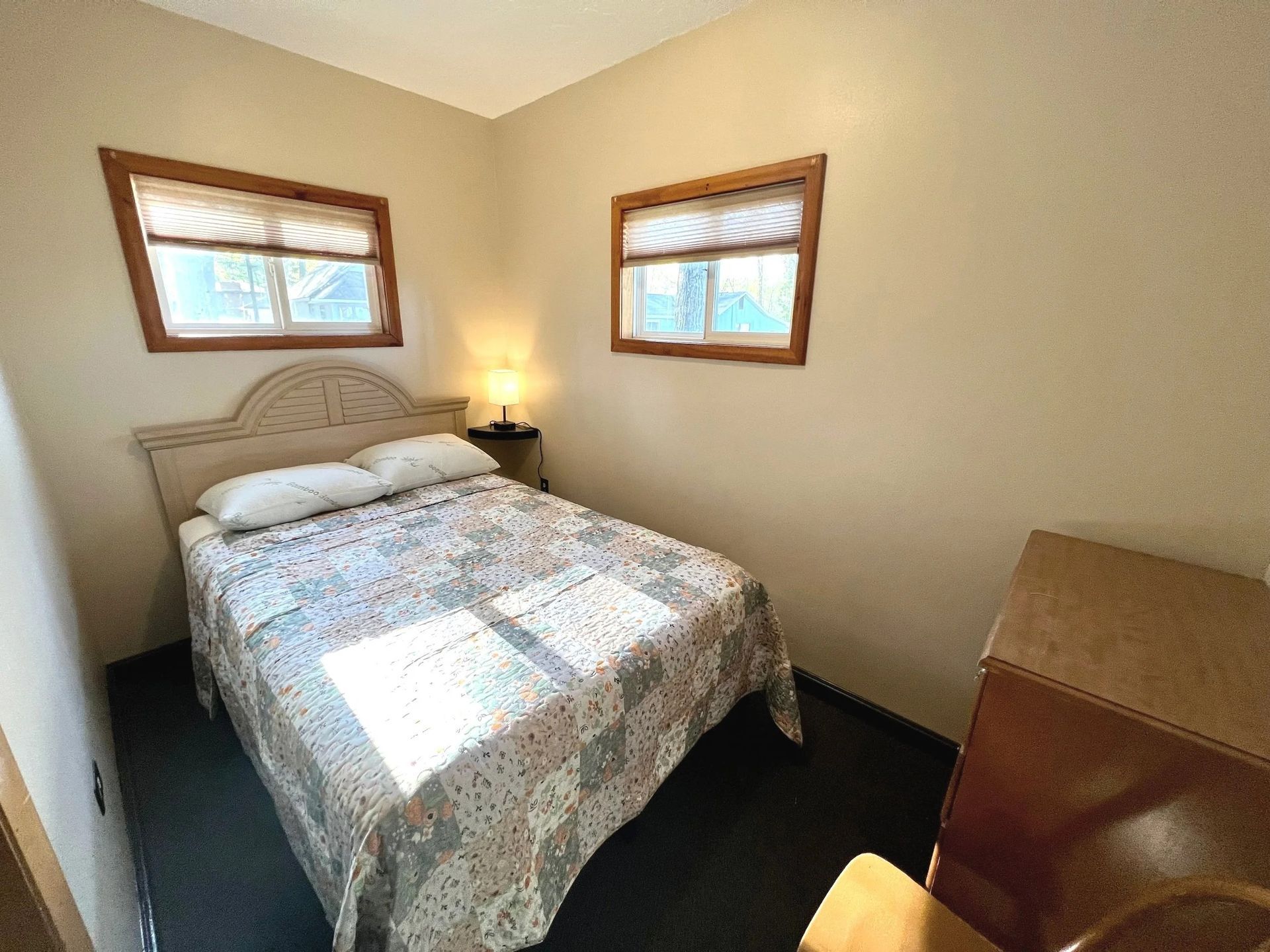 Small bedroom with a bed, two windows, nightstand with lamp, and a dresser.