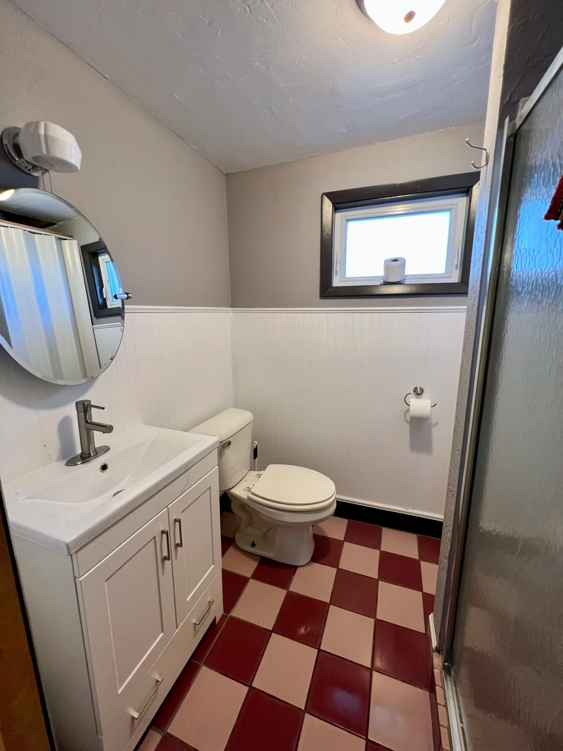 Bathroom with white vanity and toilet, red and white checkered floor, small window, and shower stall.