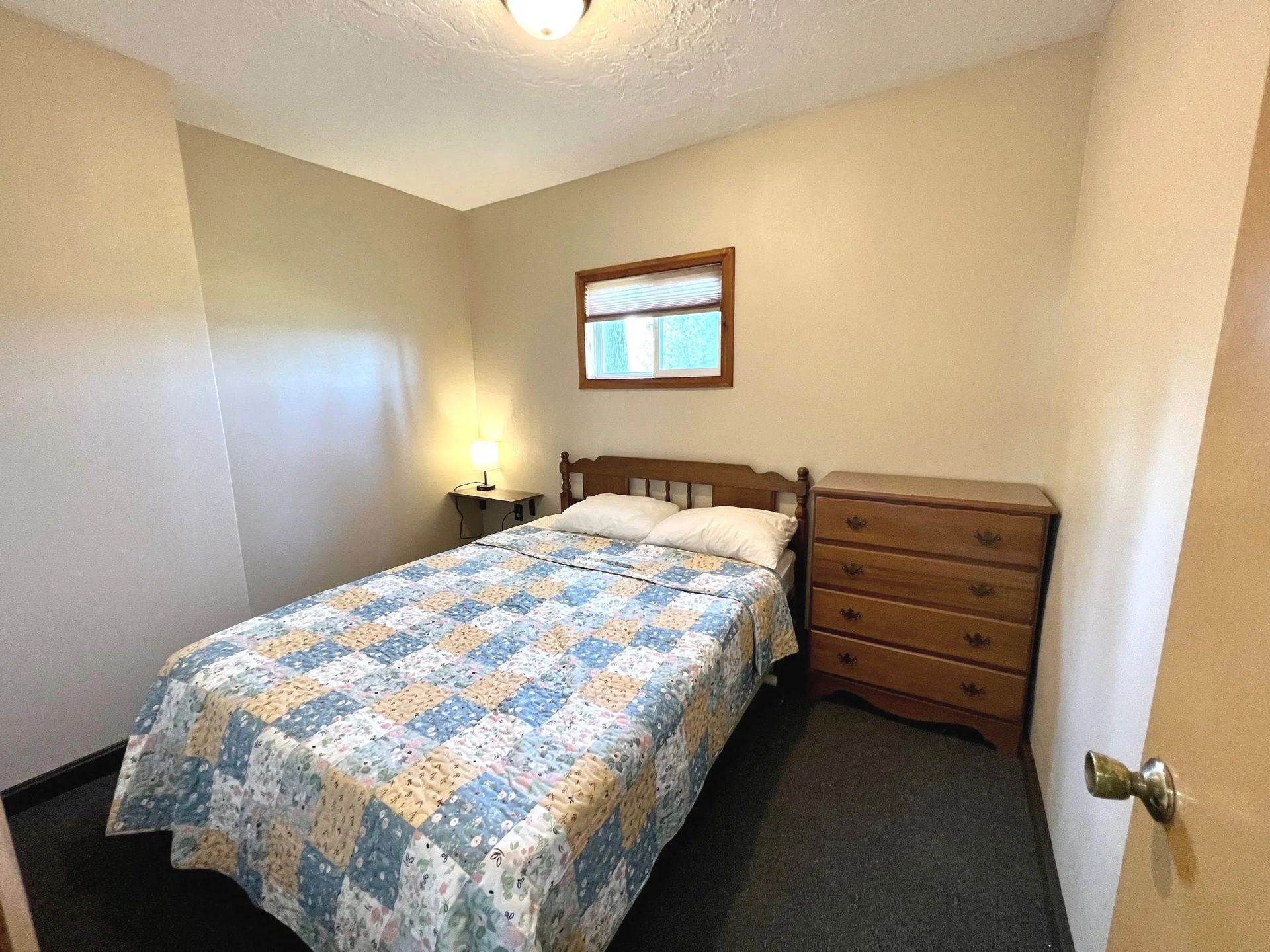 Bedroom with a double bed, dresser, small window, and a bedside table with a lamp.