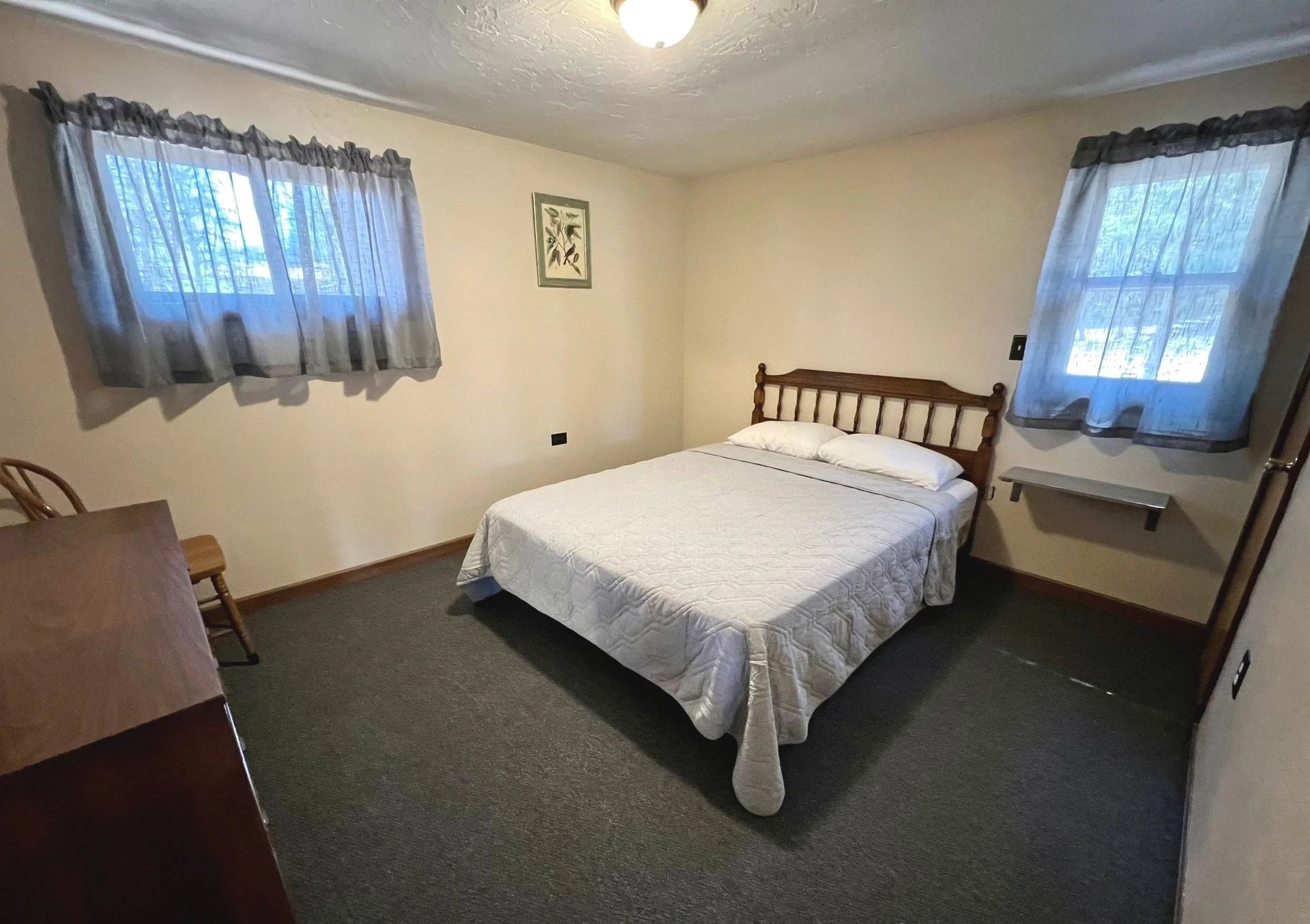Bedroom with a double bed, two windows with curtains, wooden headboard, and dresser.
