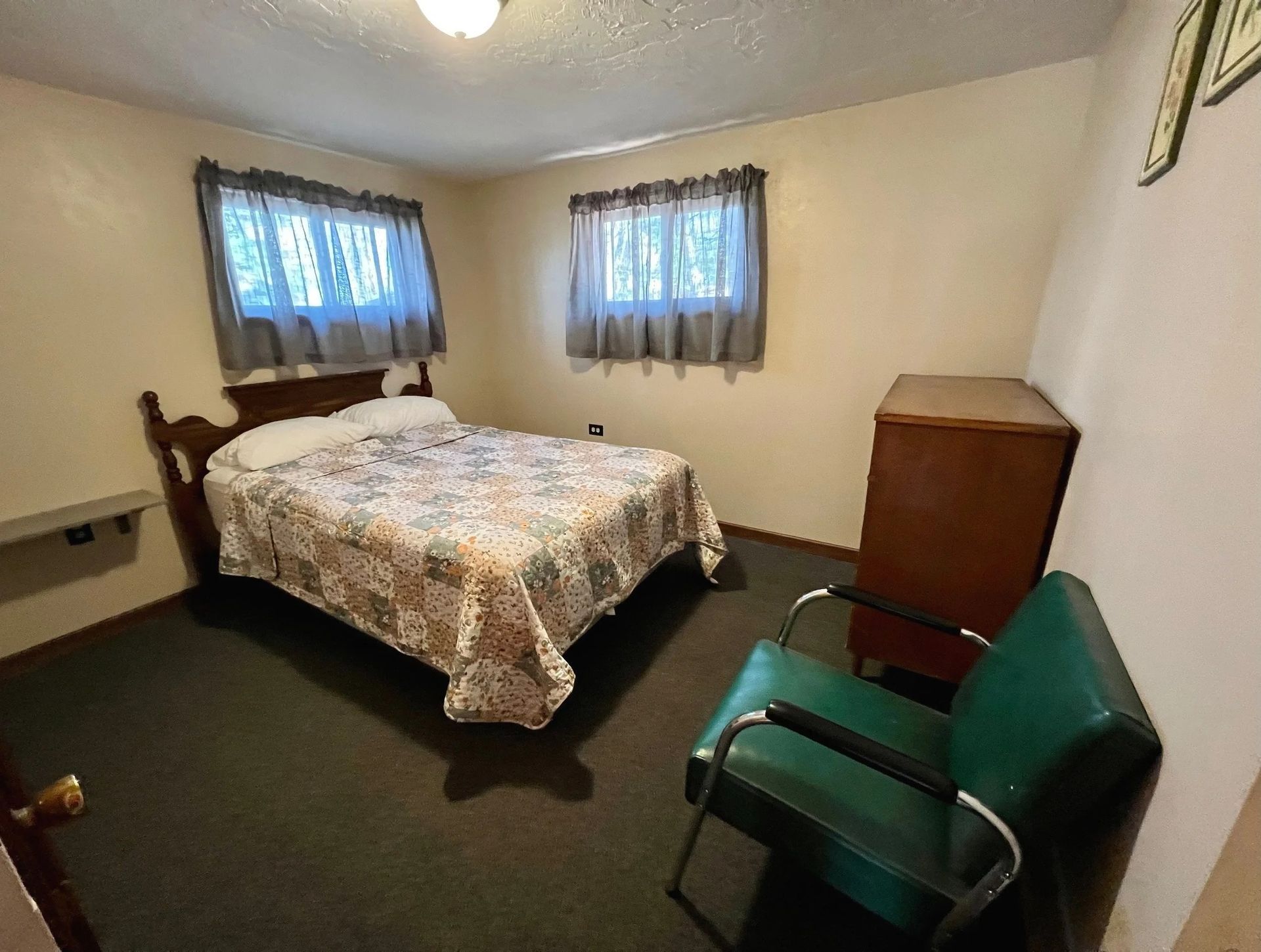 Bedroom with bed, dresser, and green chair. Windows with curtains and dark carpet.