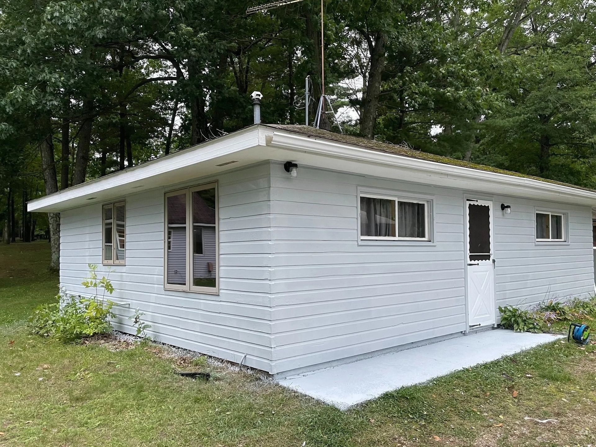 Small, light-blue cottage with windows and a gray patio in front of trees.