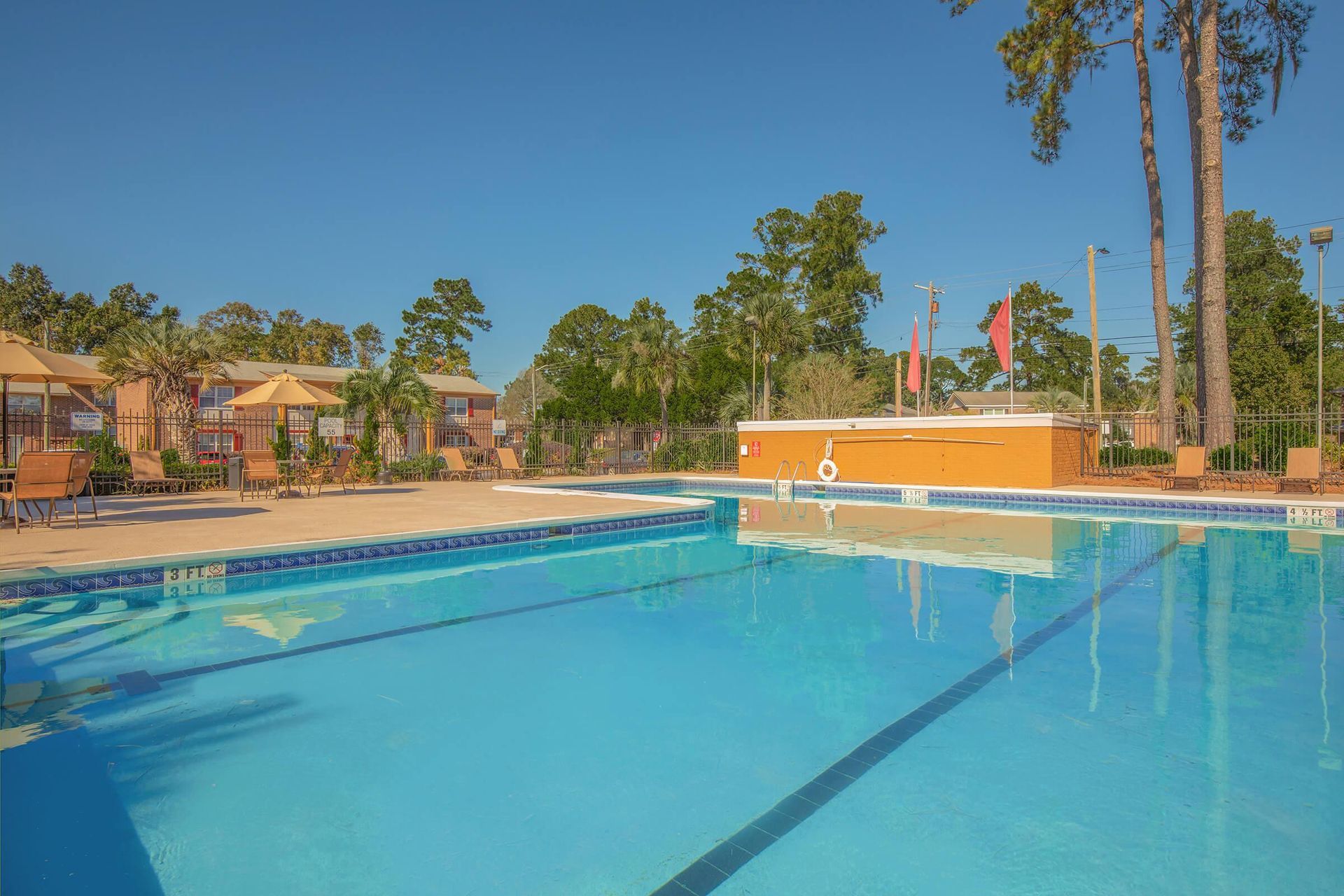 Outdoor swimming pool with blue water. Buildings and trees in the background under a clear sky.