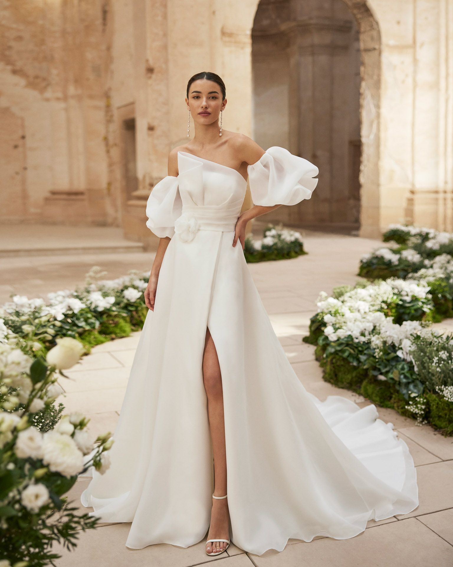 Donna in abito da sposa bianco con maniche a sbuffo monospalla e spacco profondo, in posa all'aperto.