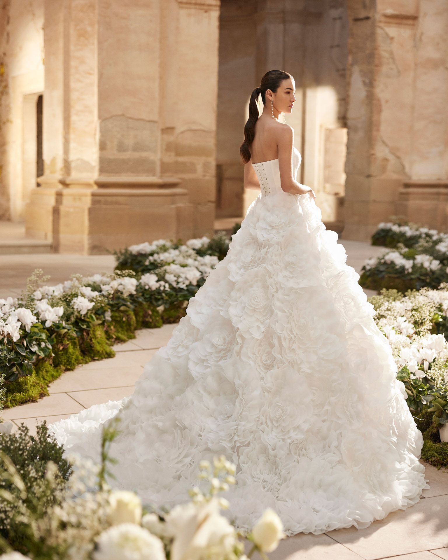 Sposa in abito bianco senza spalline con volant e lungo strascico, in piedi all'aperto. Fiori bianchi e colonne di pietra.