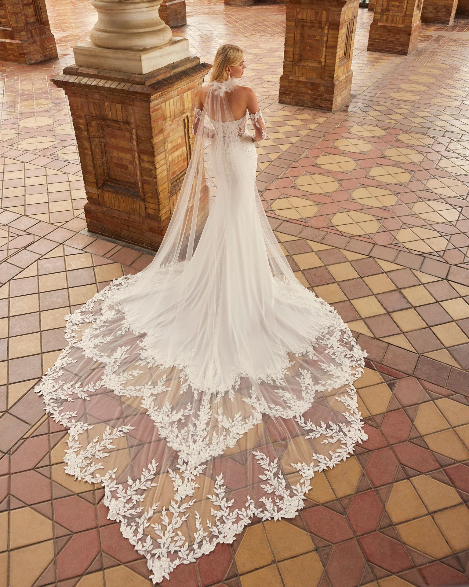 Sposa bionda in abito da sposa bianco a sirena con lungo strascico, in posa su un pavimento decorato.