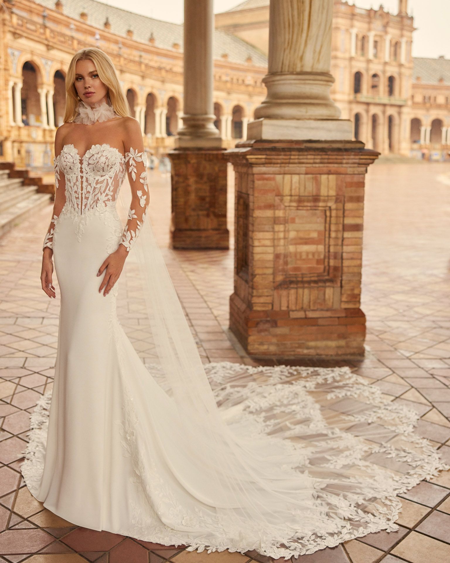 Donna bionda in abito da sposa bianco con lungo strascico, in piedi all'aperto vicino ai pilastri.