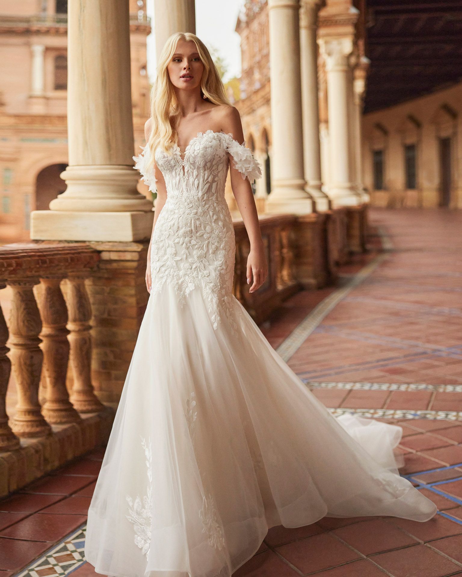 Una donna bionda con un abito da sposa a spalle scoperte posa all'aperto su un vialetto di mattoni.