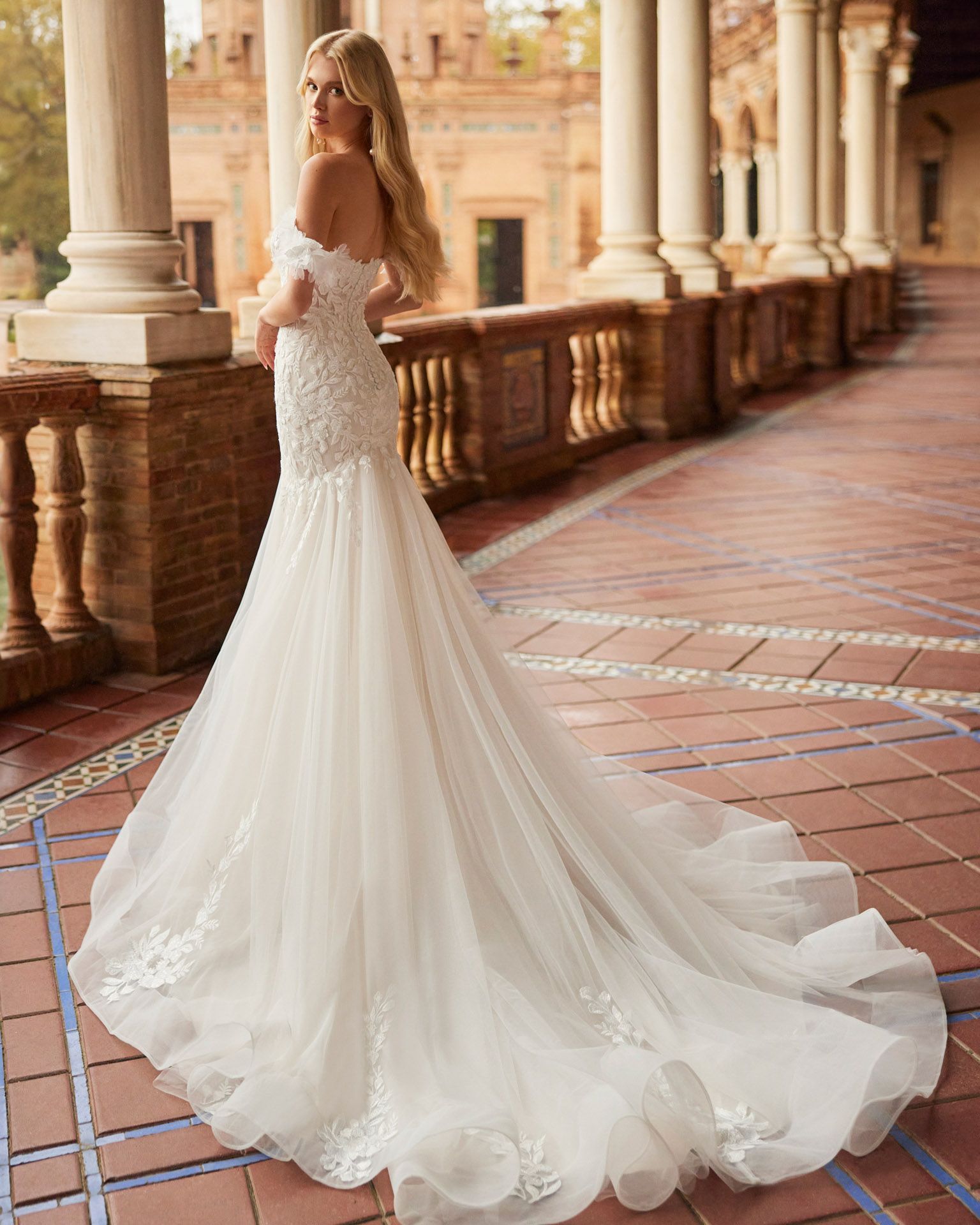 Una donna bionda, con un abito da sposa bianco a spalle scoperte e un lungo strascico, è in piedi su un balcone piastrellato.