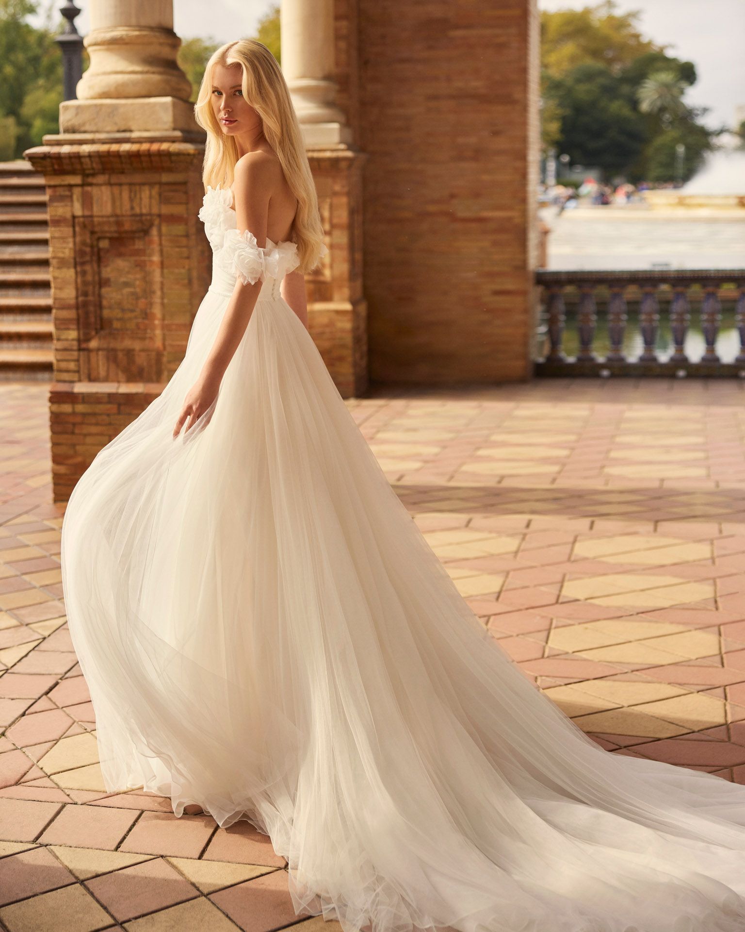 Donna bionda in abito da sposa bianco con spalle scoperte, che cammina all'aperto.