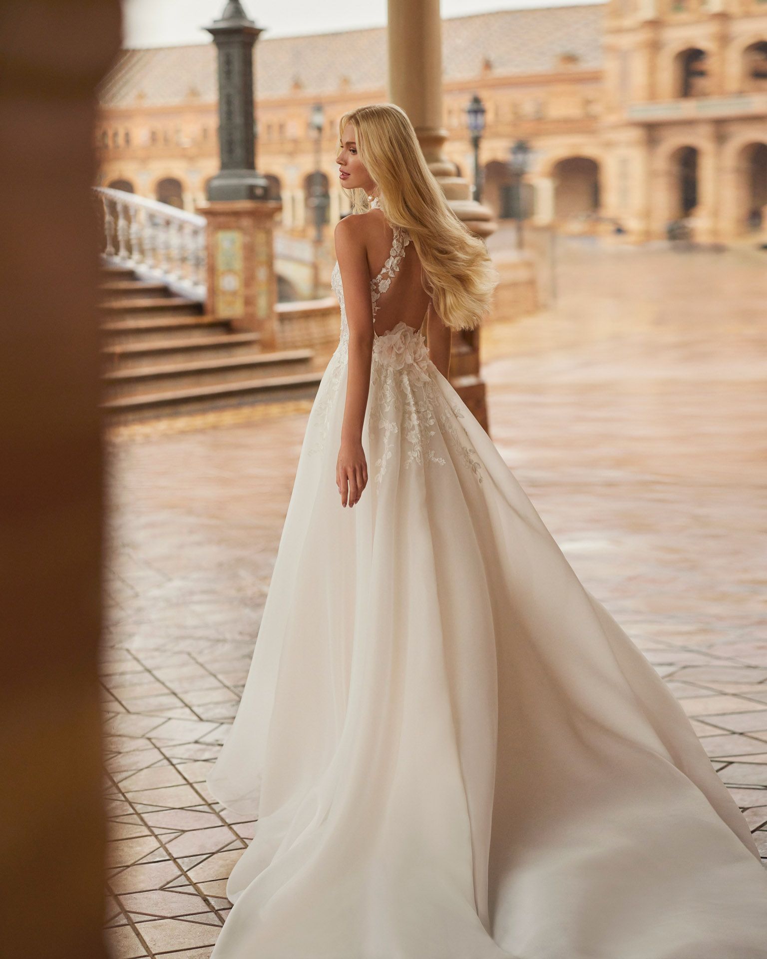 Una donna bionda in abito da sposa con strascico posa su un marciapiede piastrellato.