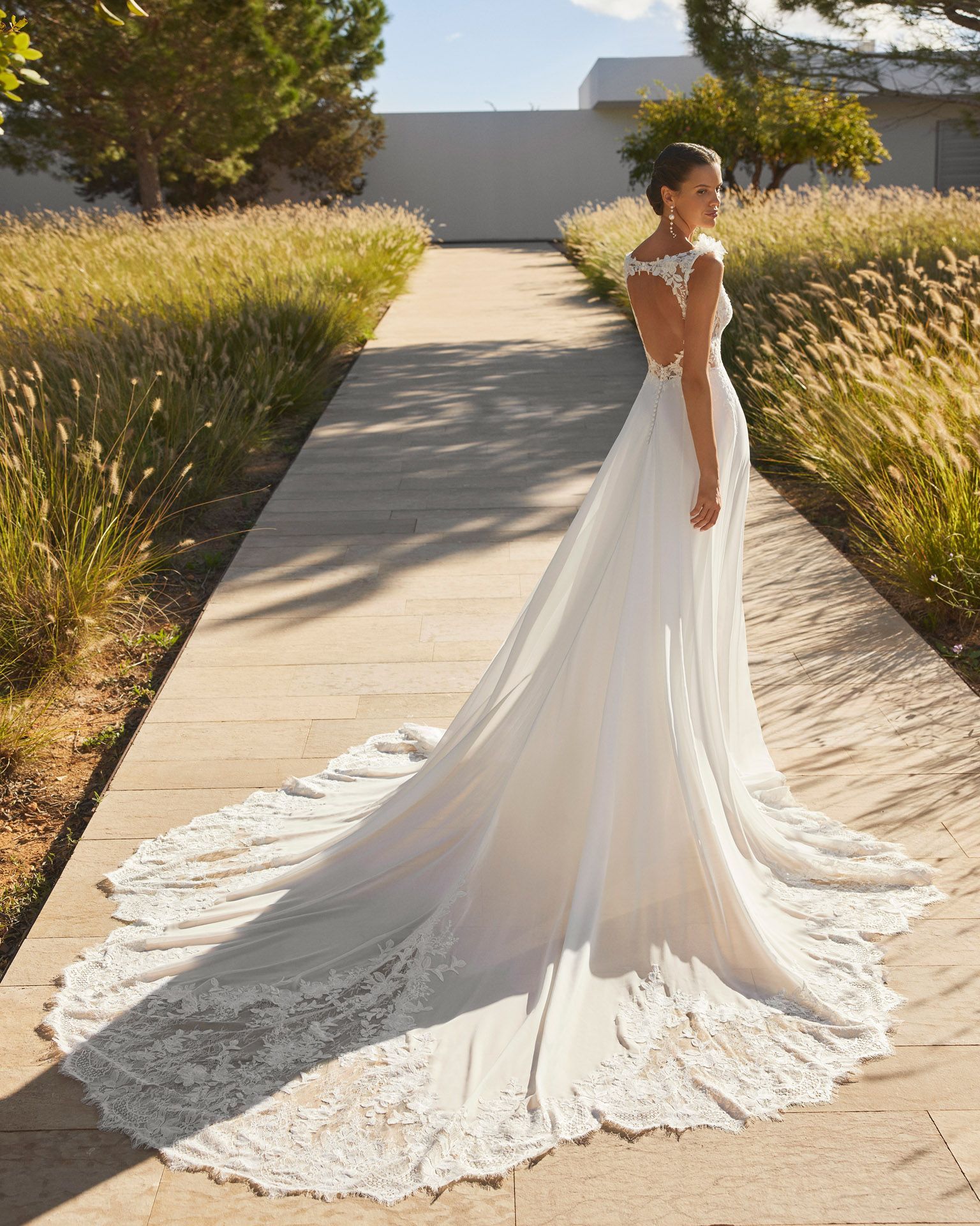 Sposa in abito da sposa bianco con lungo strascico, in piedi su un sentiero di pietra all'aperto.