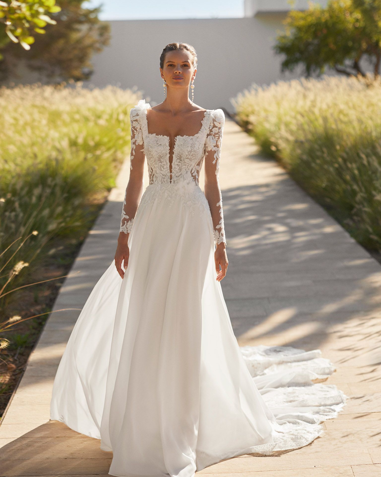 Donna in abito da sposa bianco con maniche in pizzo e strascico, in piedi all'aperto.