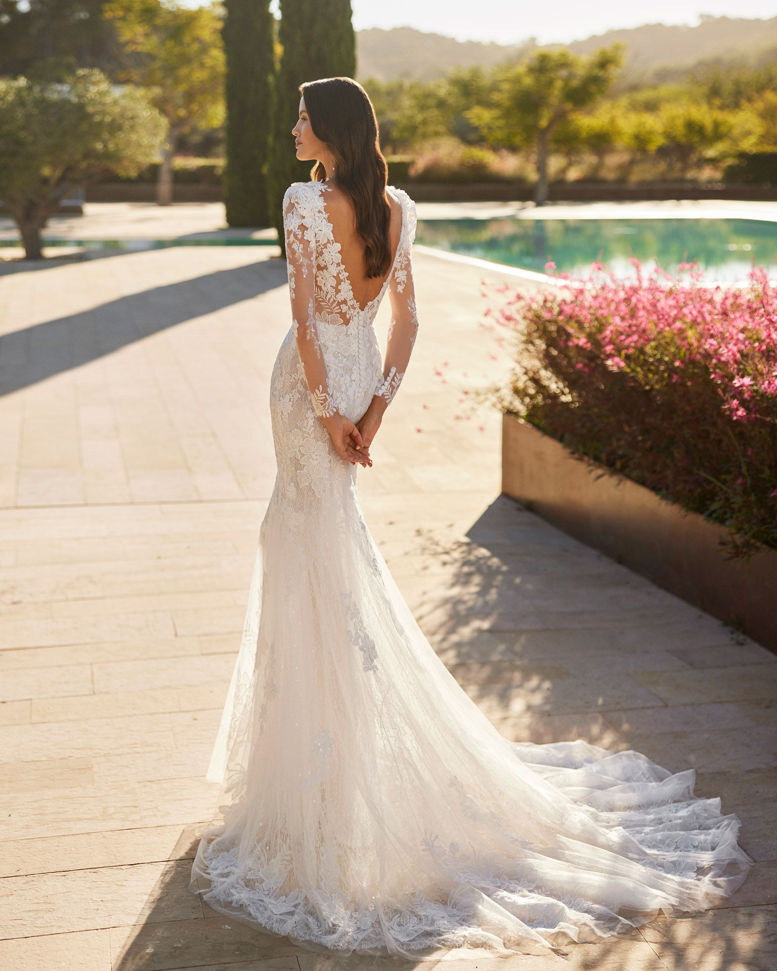 Donna in abito da sposa in pizzo bianco, schiena scoperta, in piedi all'aperto con piscina, giornata di sole.