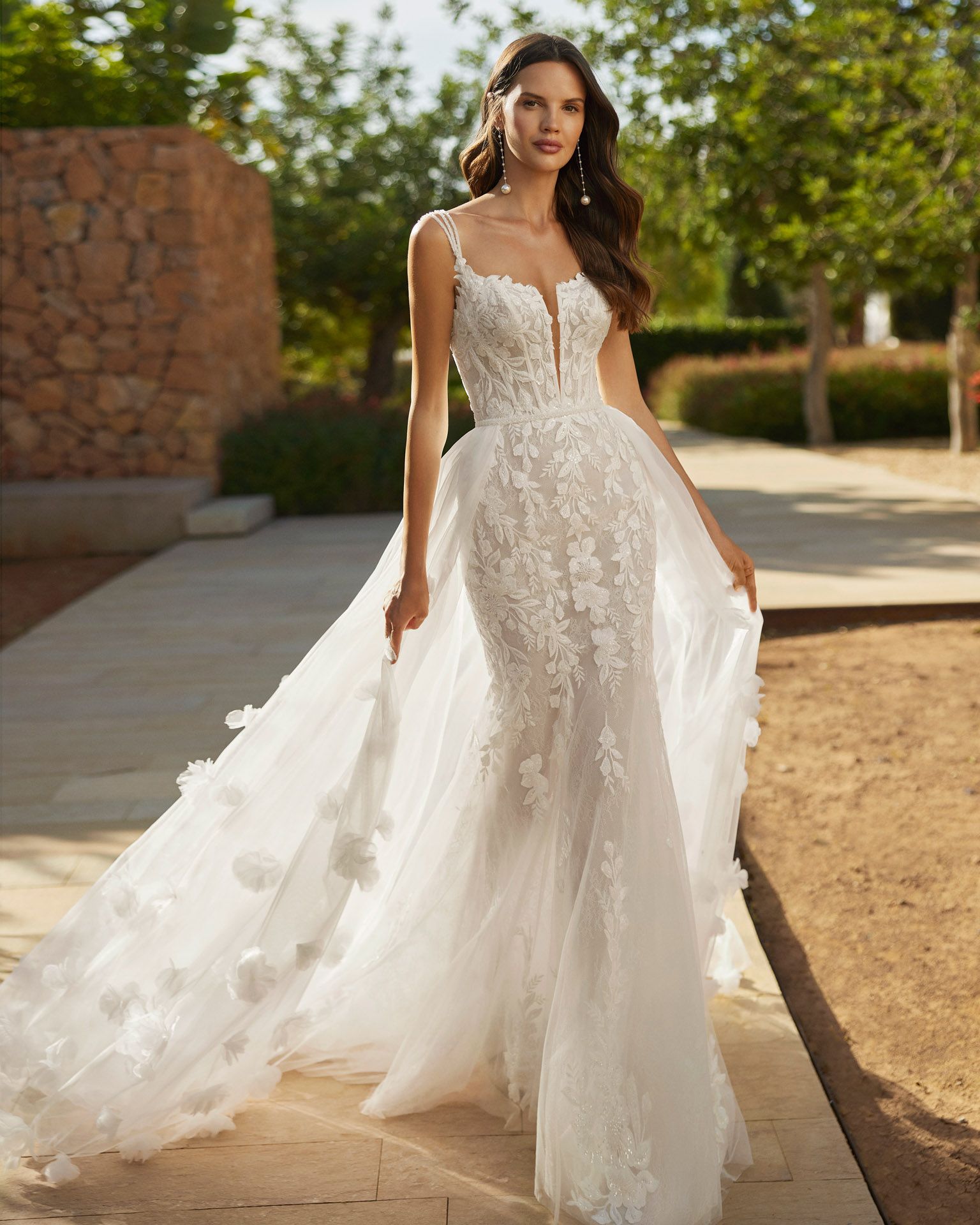 Donna in abito da sposa bianco con dettagli floreali e lungo strascico, in piedi all'aperto.