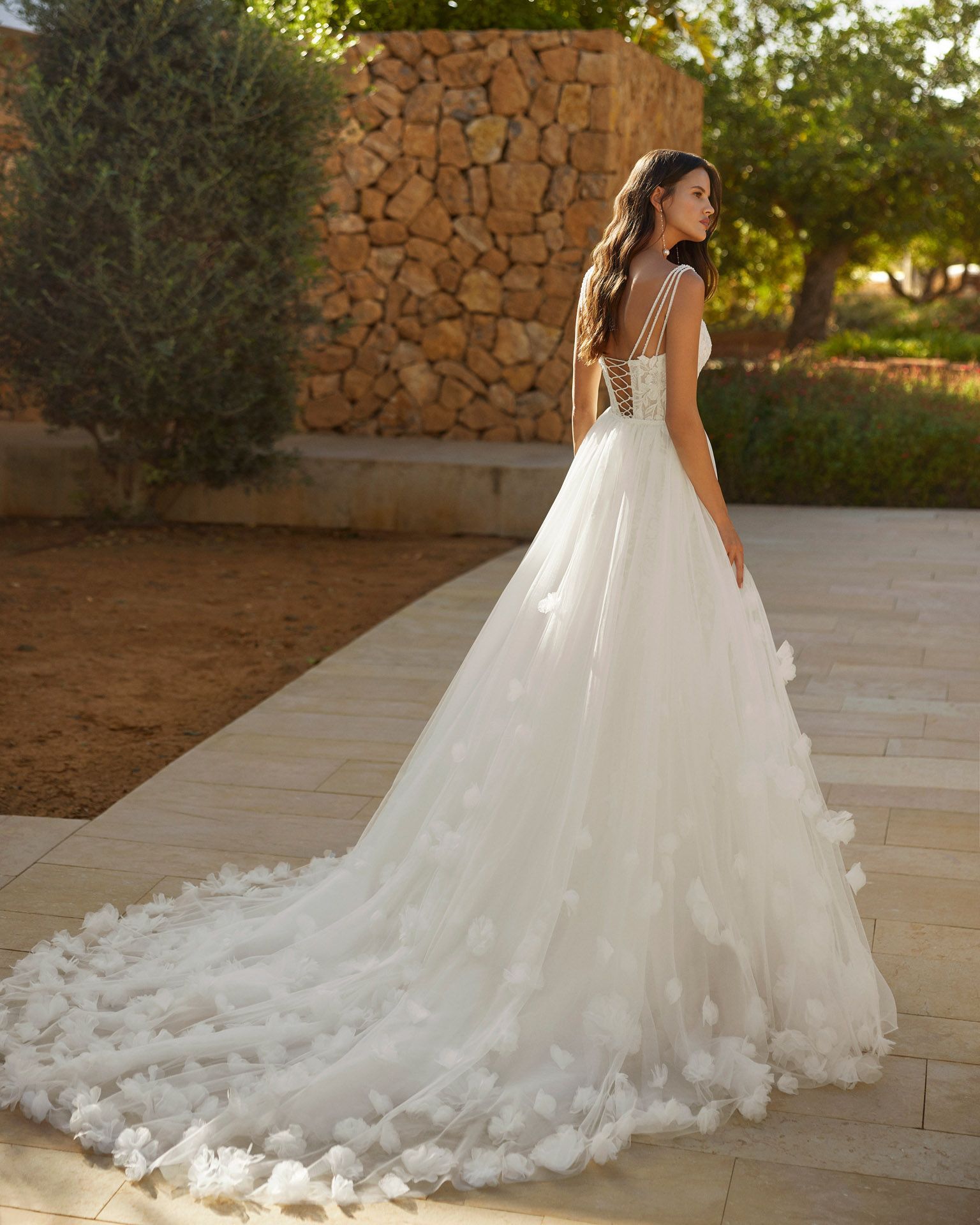 Donna in abito da sposa bianco con dettagli floreali e lungo strascico, all'aperto vicino a un muro di pietra.