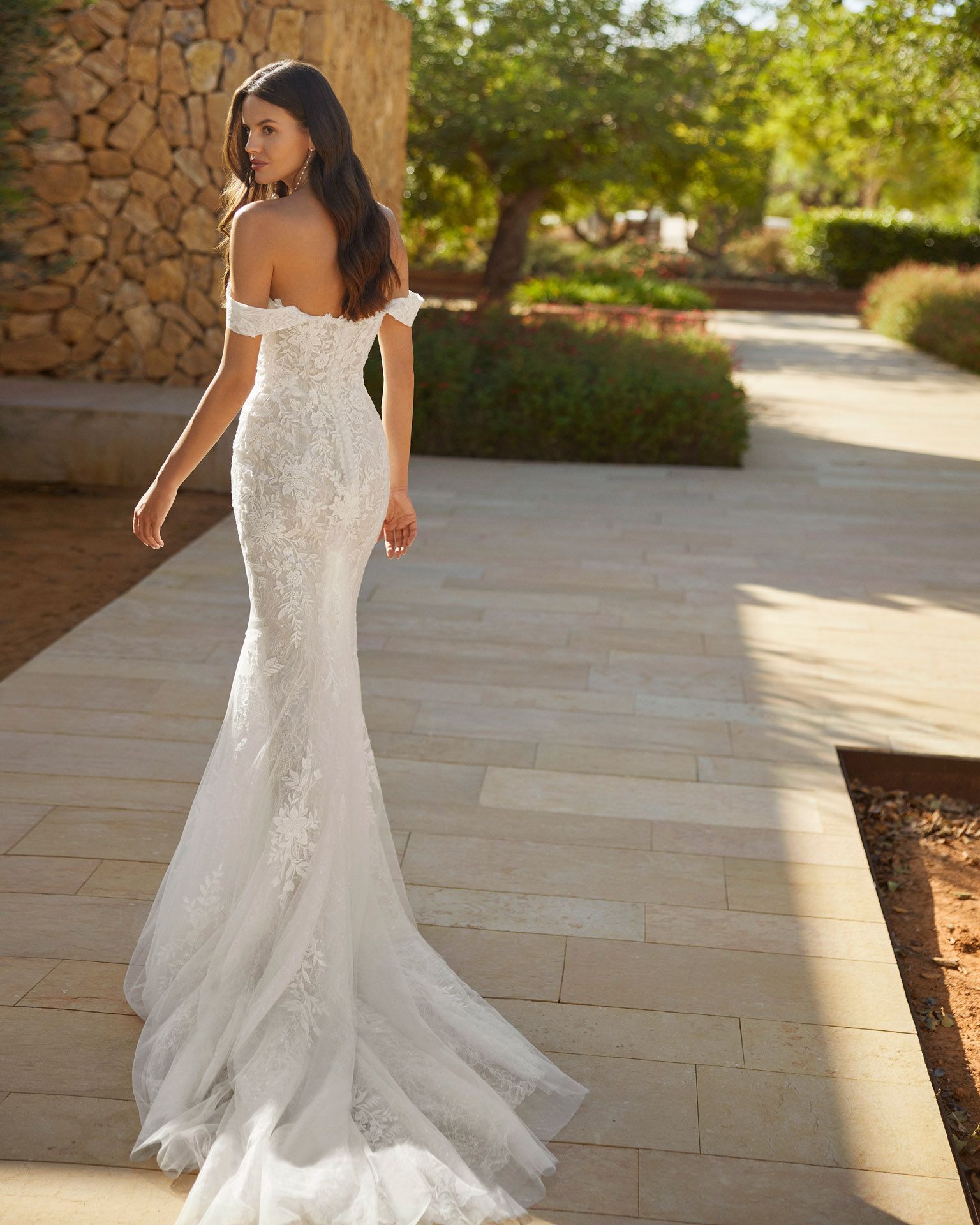 Donna in abito da sposa in pizzo bianco, con spalle scoperte e strascico, che cammina all'aperto su un sentiero lastricato.