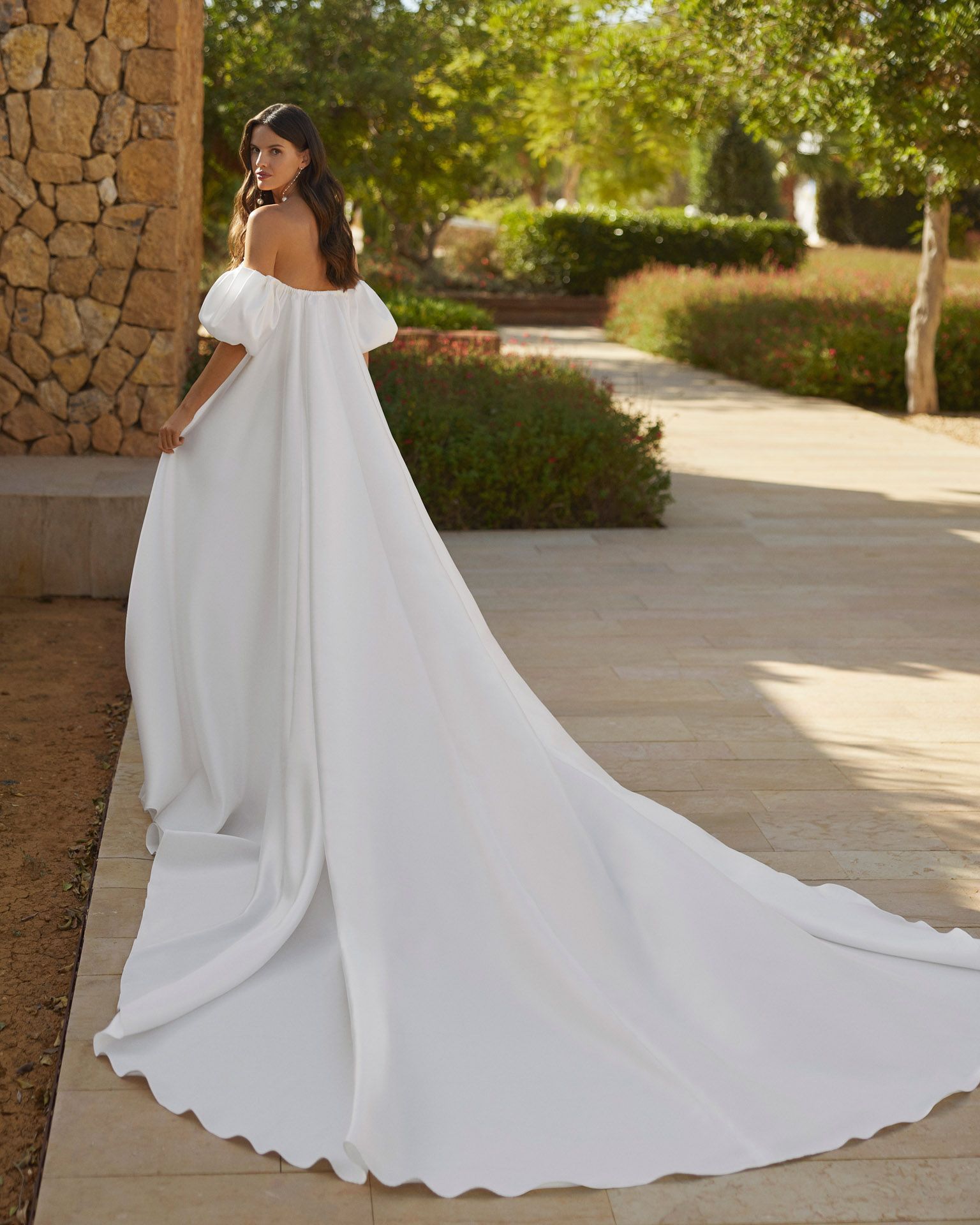 Donna in abito da sposa bianco con spalle scoperte e lungo strascico, in piedi all'aperto.